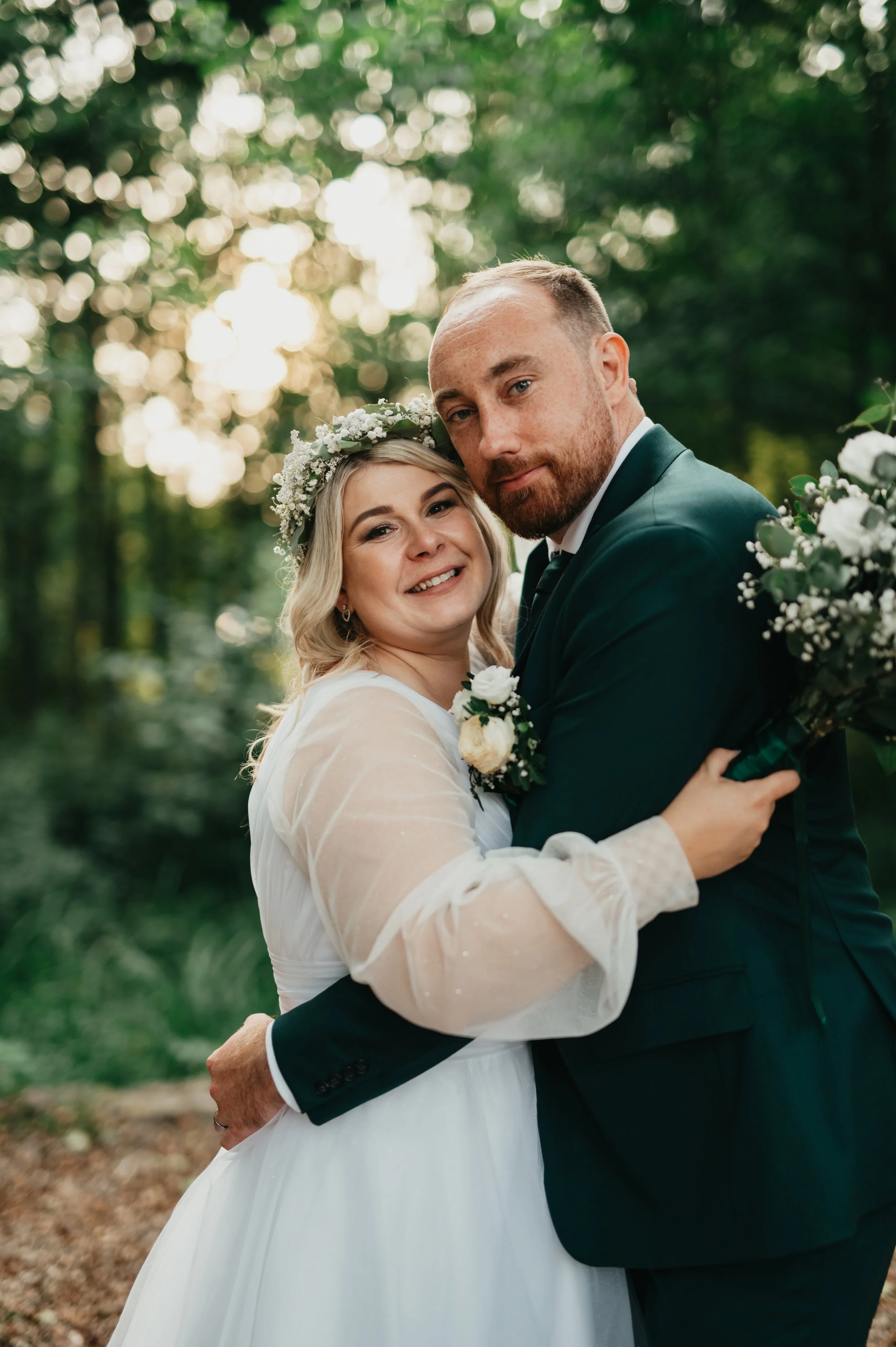 Nevěsta a ženich se objímají venku v zalesněném prostředí a usmívají se do fotoaparátu. Nevěsta má na sobě bílé svatební šaty a květinovou čelenku; ženich má na sobě tmavě zelený oblek.