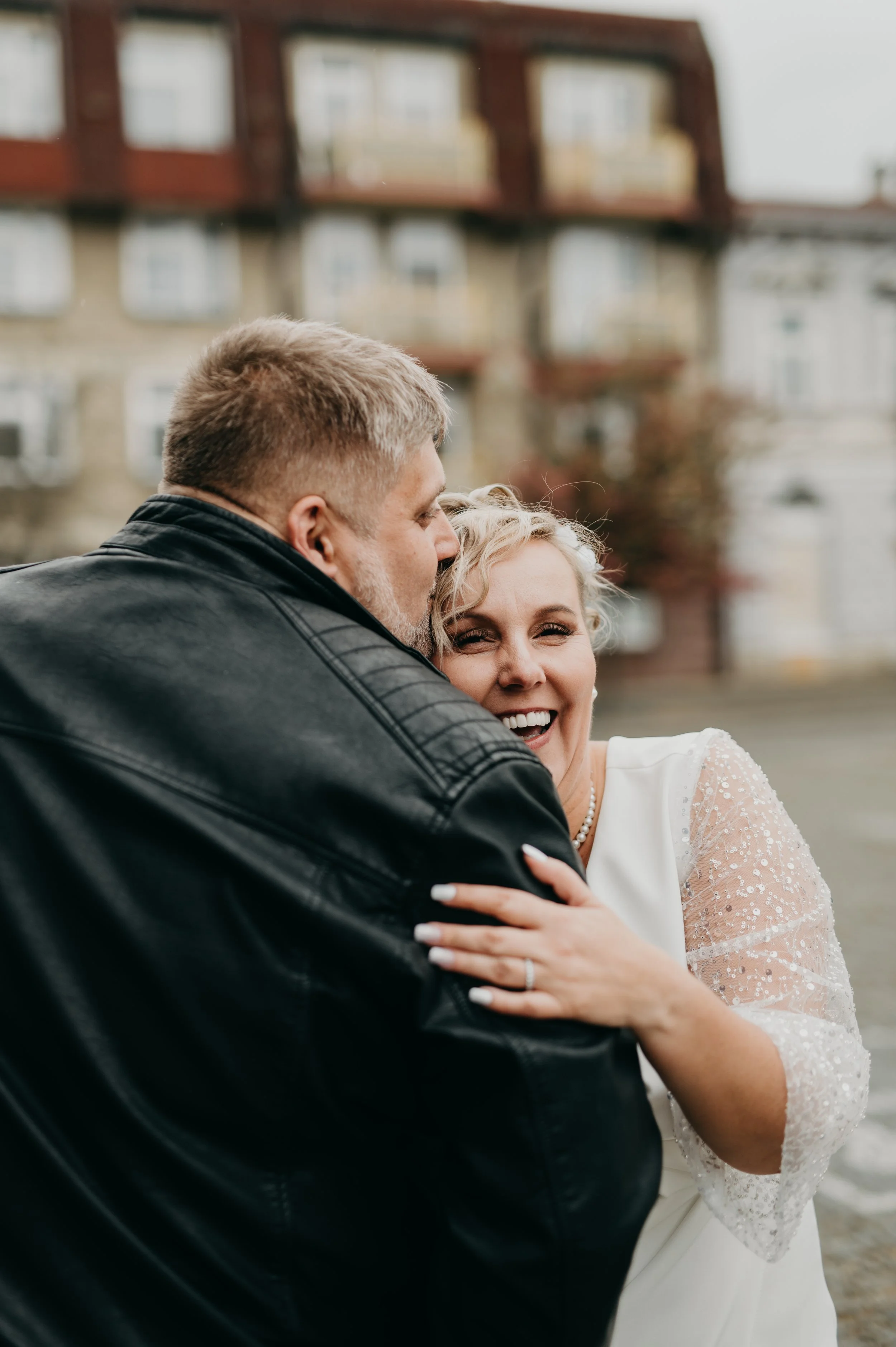 A joyful woman with blonde curly hair wearing a white dress with sheer lace sleeves, smiling and hugging a man in a black leather jacket outdoors with a blurry apartment building in the background.