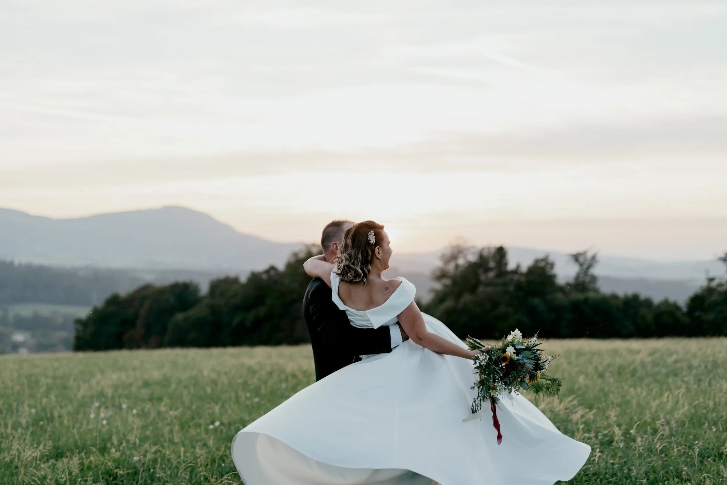 Man carrying woman in white dress with bouquet in a field at sunset, mountains in background.
