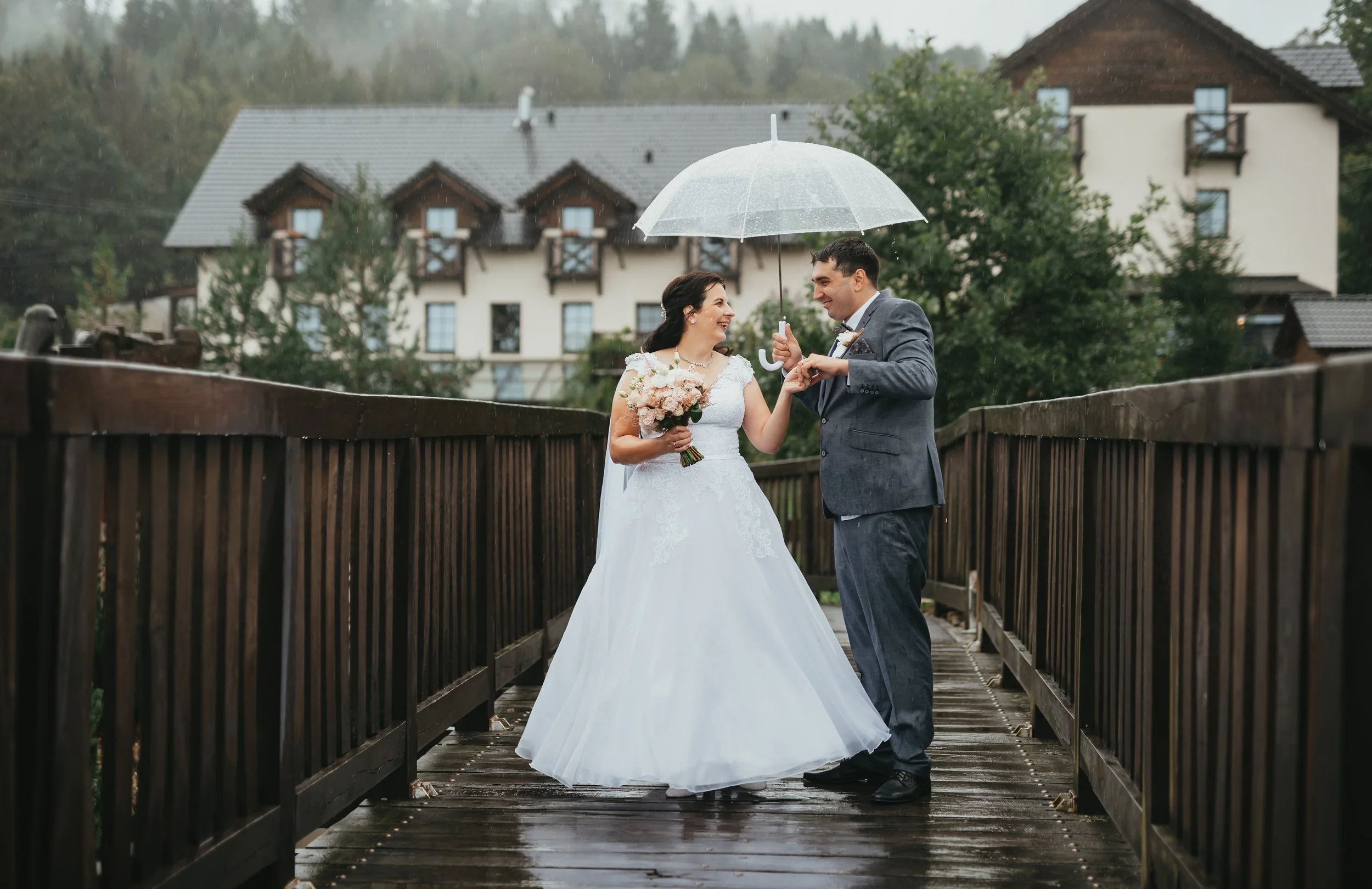 Nevěsta a ženich stojící na dřevěném mostě v dešti, držící deštník a usmívající se na sebe, s domem a stromy v pozadí.
