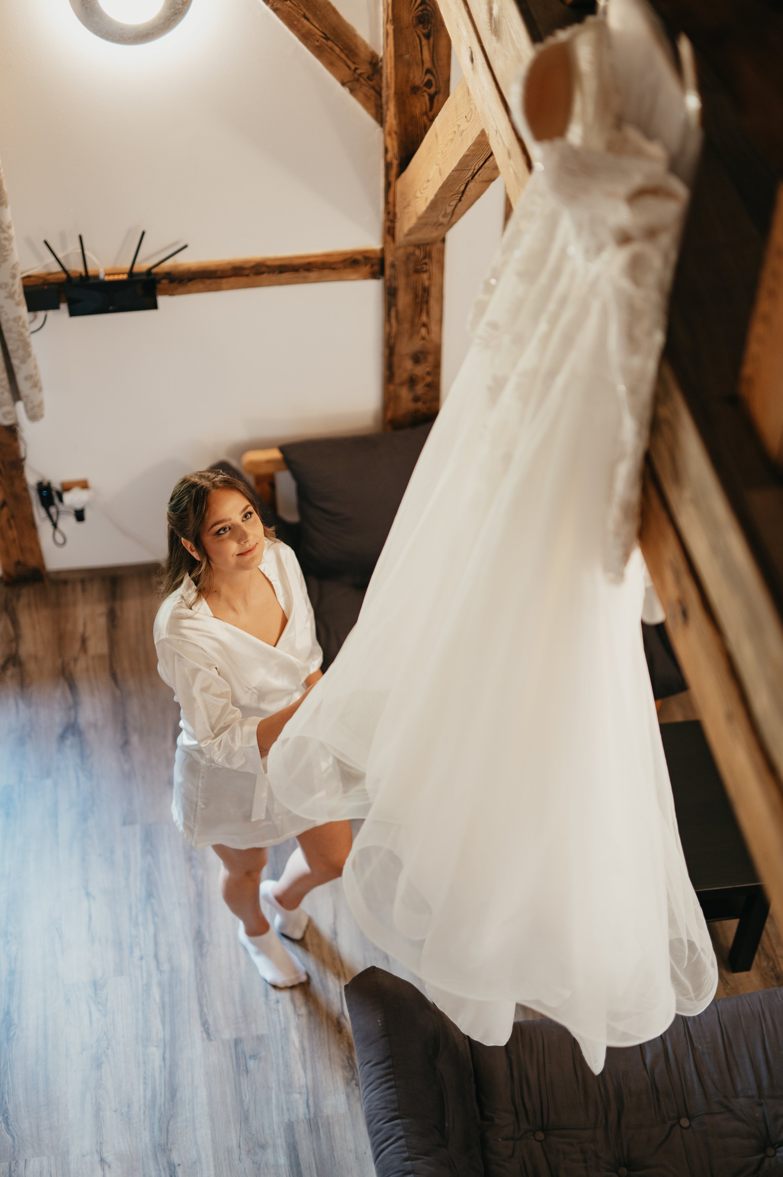 A woman in a satin robe looks up at a wedding dress hanging from a loft ceiling.