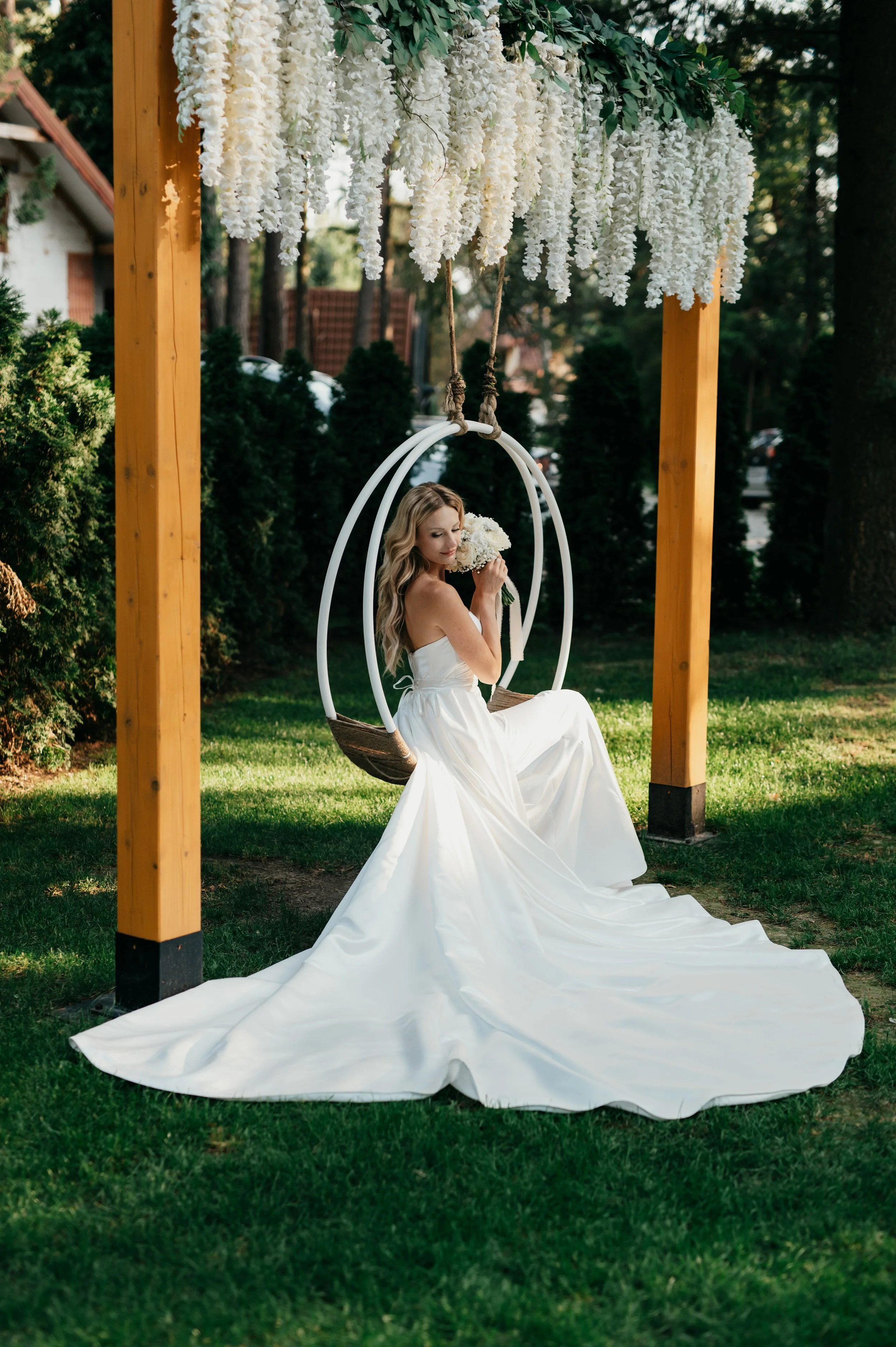 A bride in a white wedding dress sitting on a swing beneath hanging white flowers, outdoors with greenery and trees in the background.