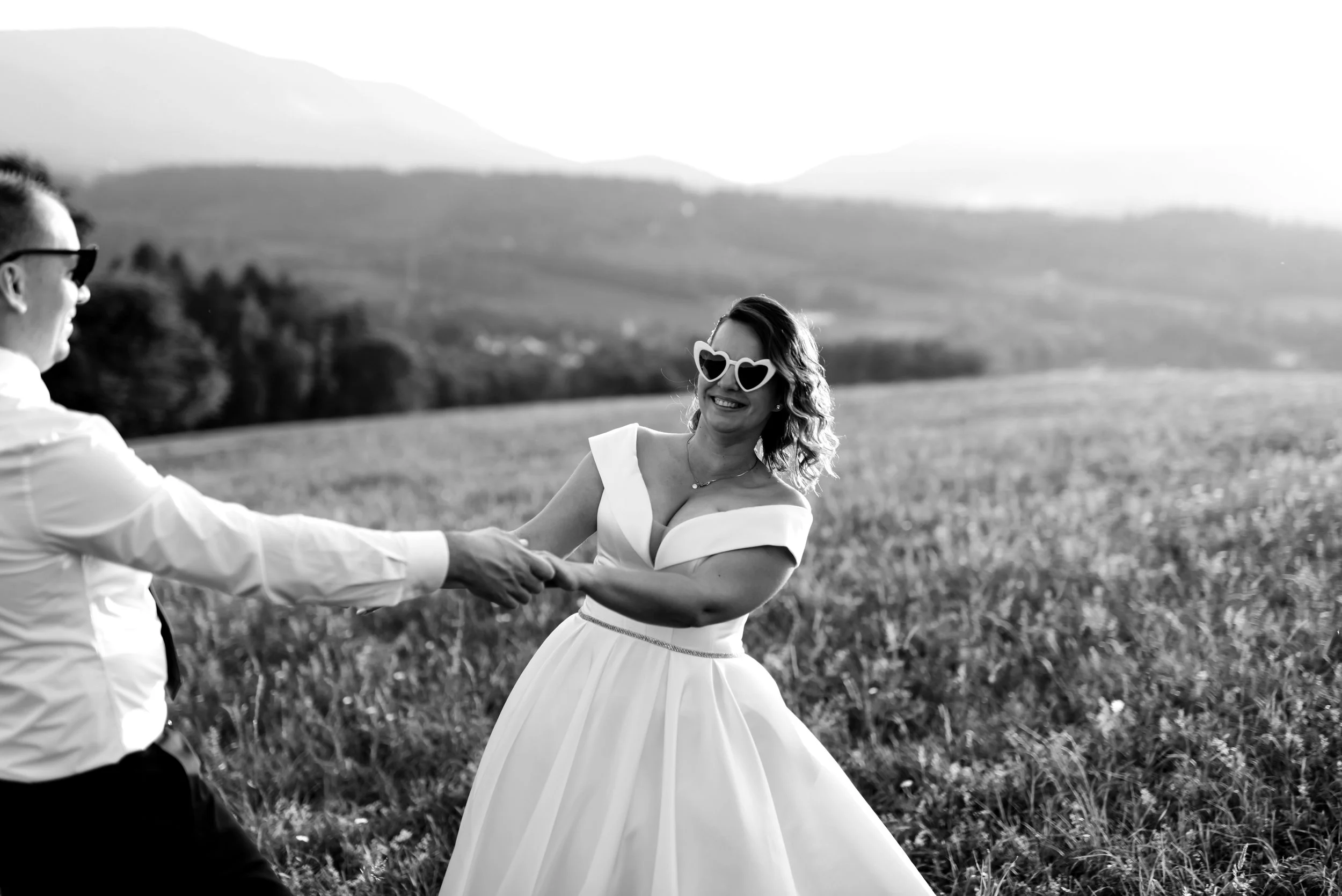 A couple holding hands and dancing in a field, woman wearing heart-shaped sunglasses and a dress, man in a shirt and tie. Black and white photo.