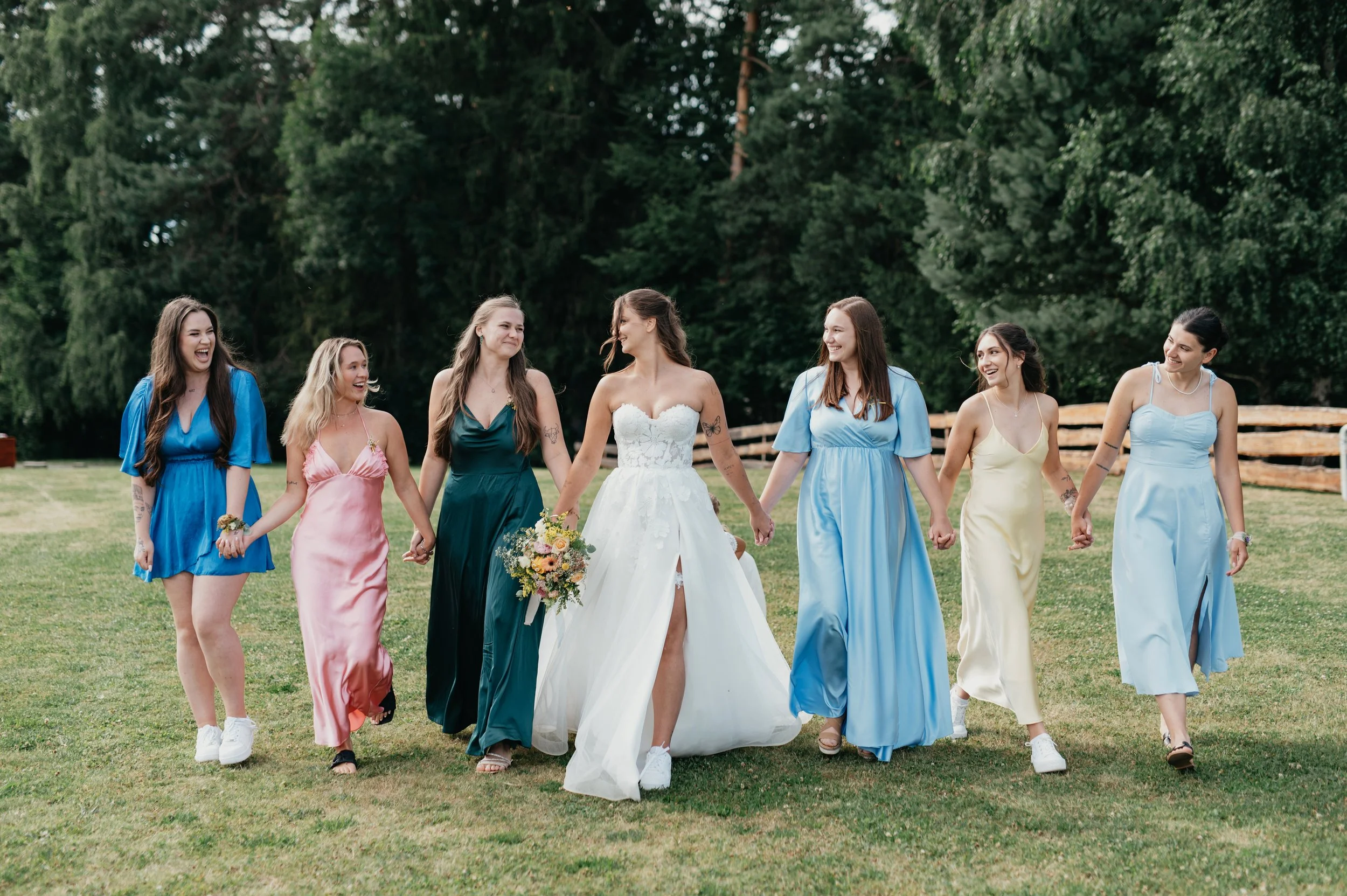 Bride in a white wedding dress holding hands with six bridesmaids in colorful dresses walking on a grassy field with trees in the background.