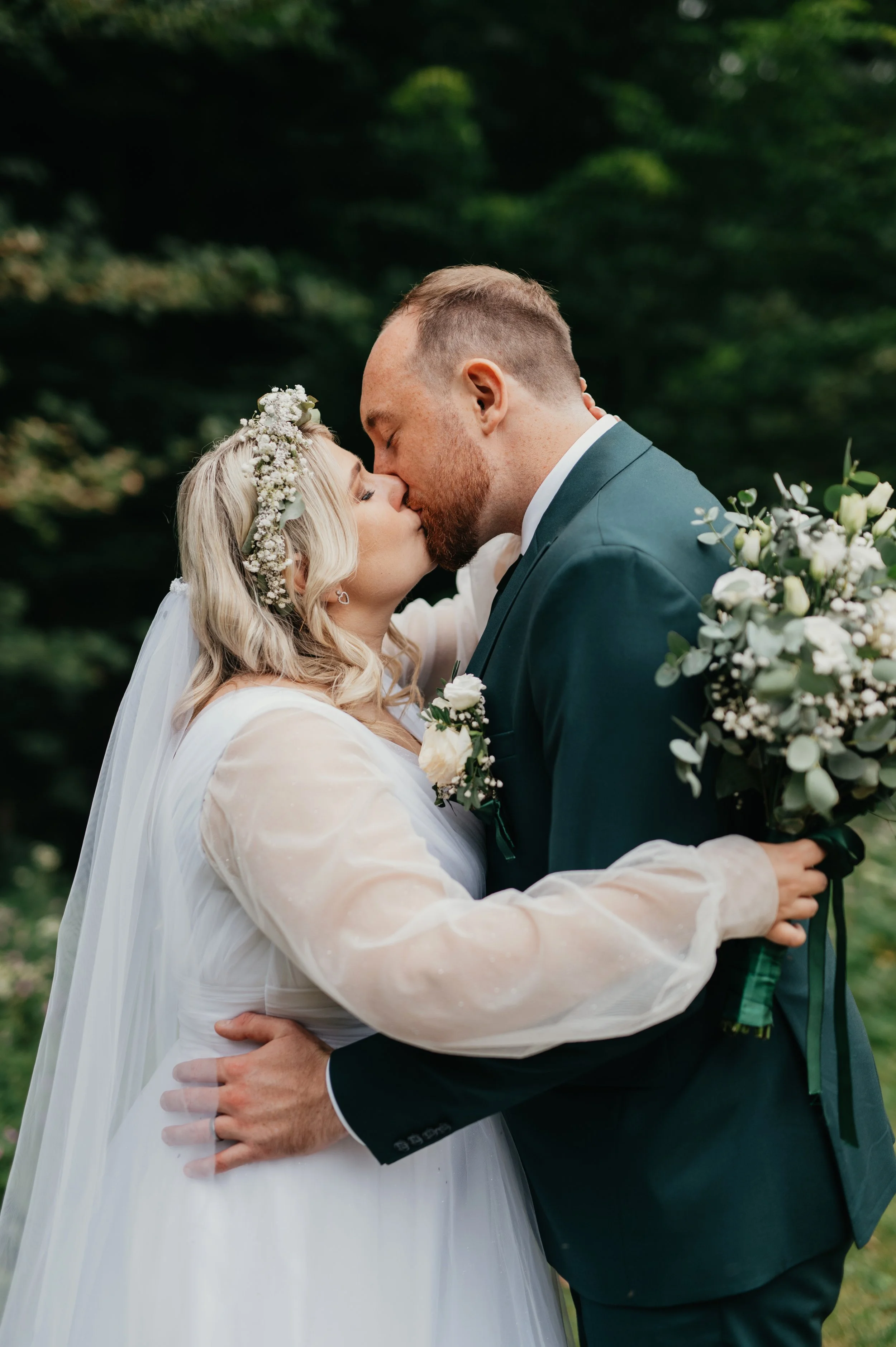 A newlywed couple sharing a kiss outdoors, with a backdrop of greenery. The bride holds a bouquet of white flowers and wears a floral crown, while the groom wears a dark suit.