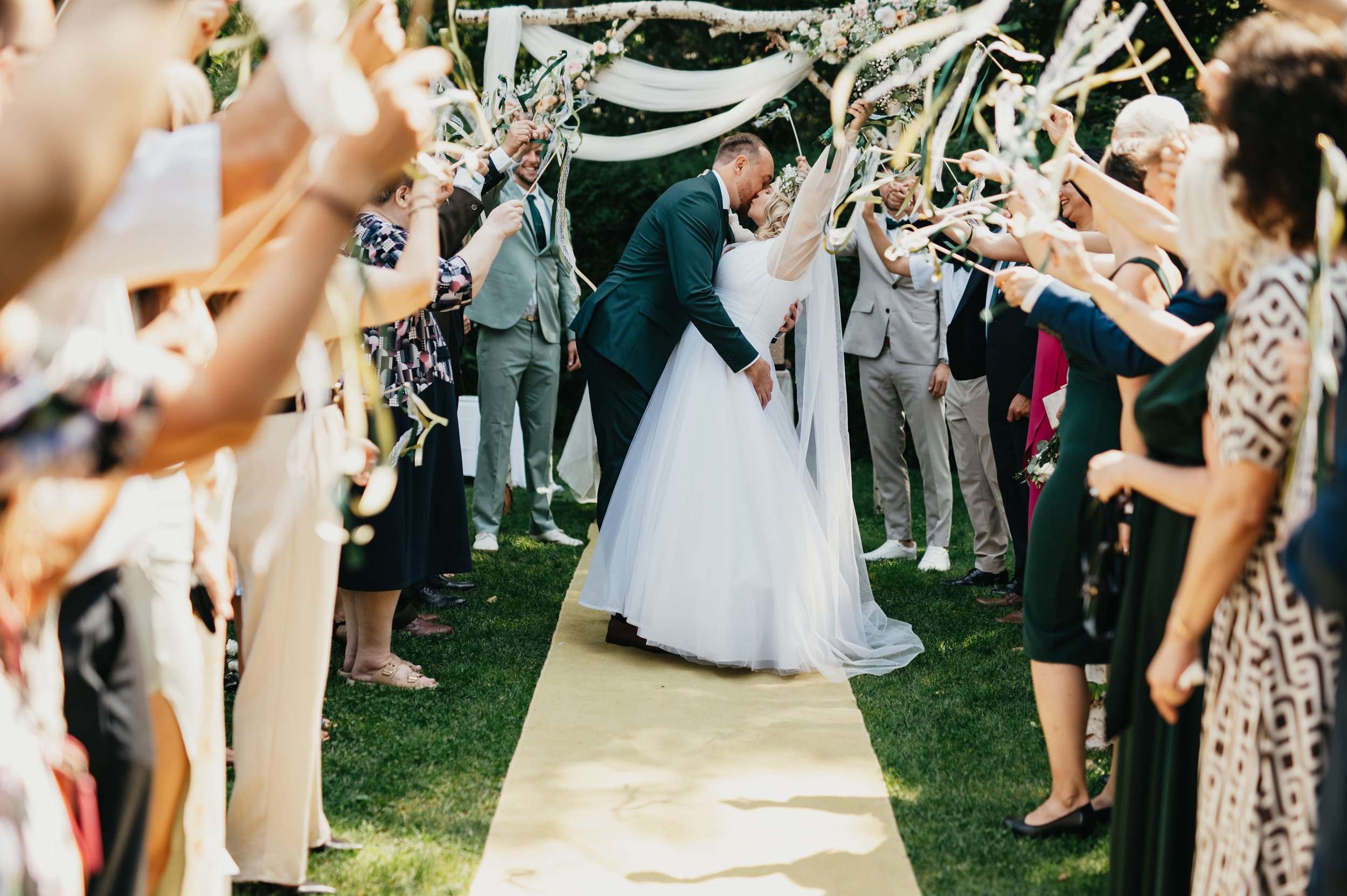 A bride and groom kiss under a floral arch at their outdoor wedding surrounded by guests holding ribbons and celebrating.