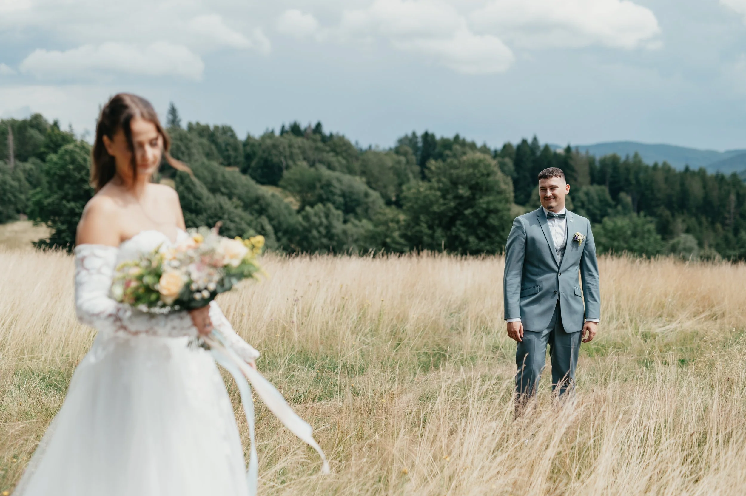 A bride holding a bouquet of flowers in a white wedding dress standing in a grassy field, with a groom in a light blue suit smiling in the distance, trees and hills in the background.