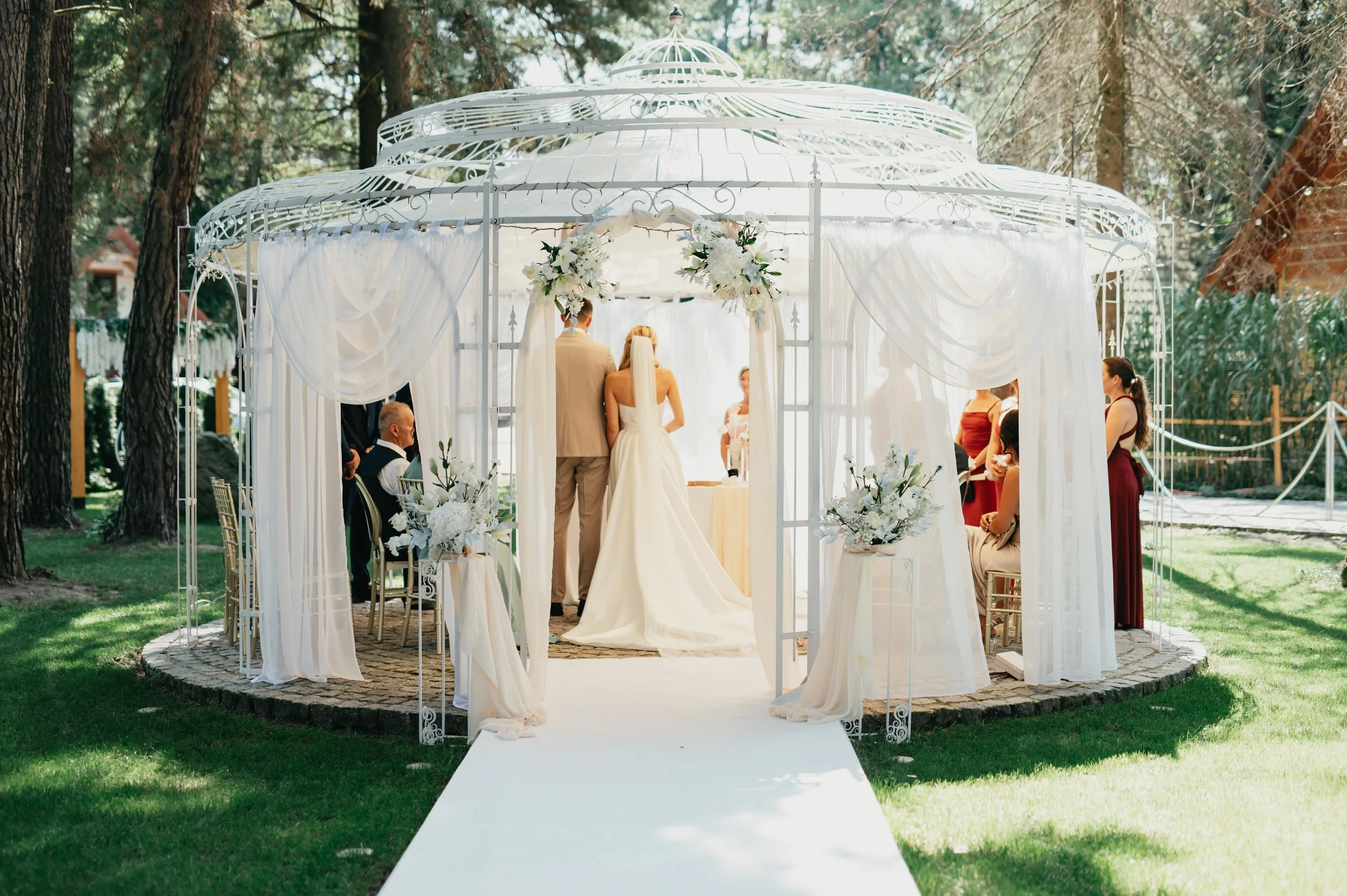 Svatební obřad v bílém altánu s bílými závěsy a květinovou výzdobou, zasazený venku mezi stromy, s nevěstou a ženichem stojícími proti sobě.