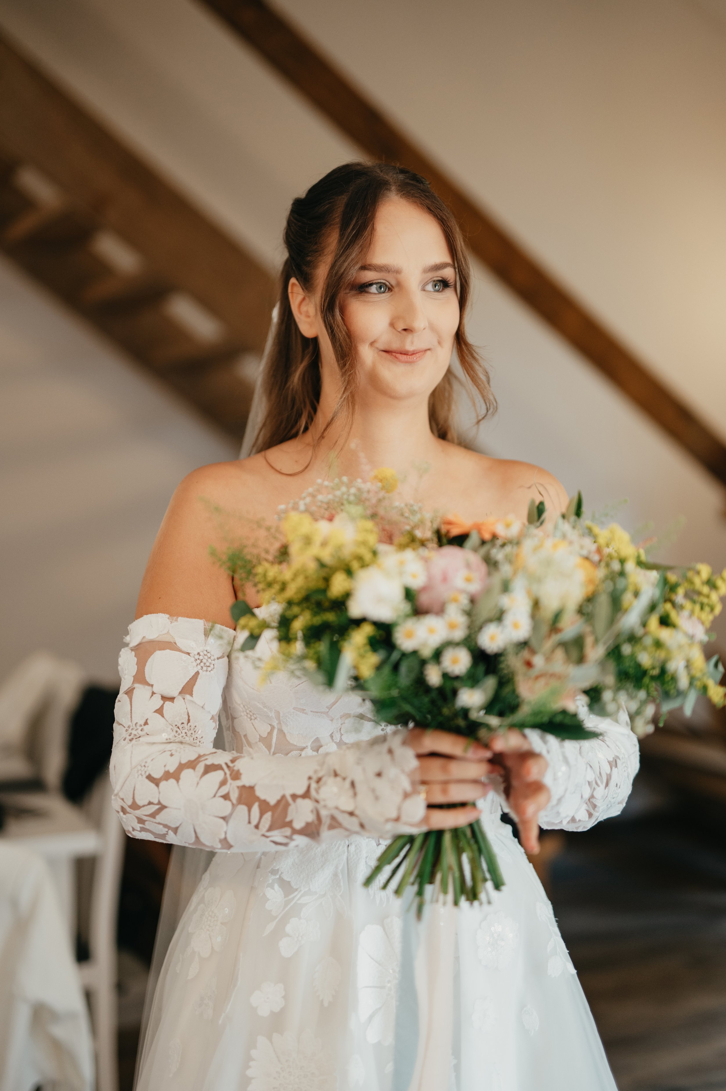 A woman in a white lace wedding dress holding a bouquet of flowers, smiling gently, indoors with a wooden beam ceiling.