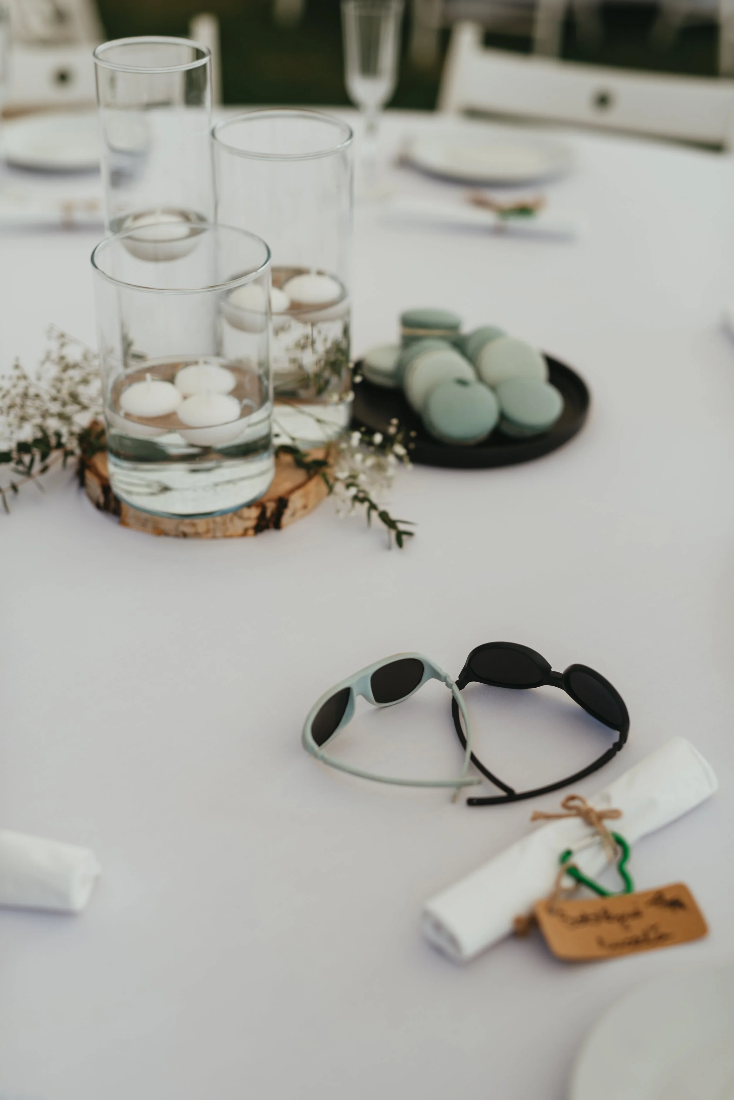 Table setting with floating candles, macarons, sunglasses, and a rolled napkin on a white tablecloth.