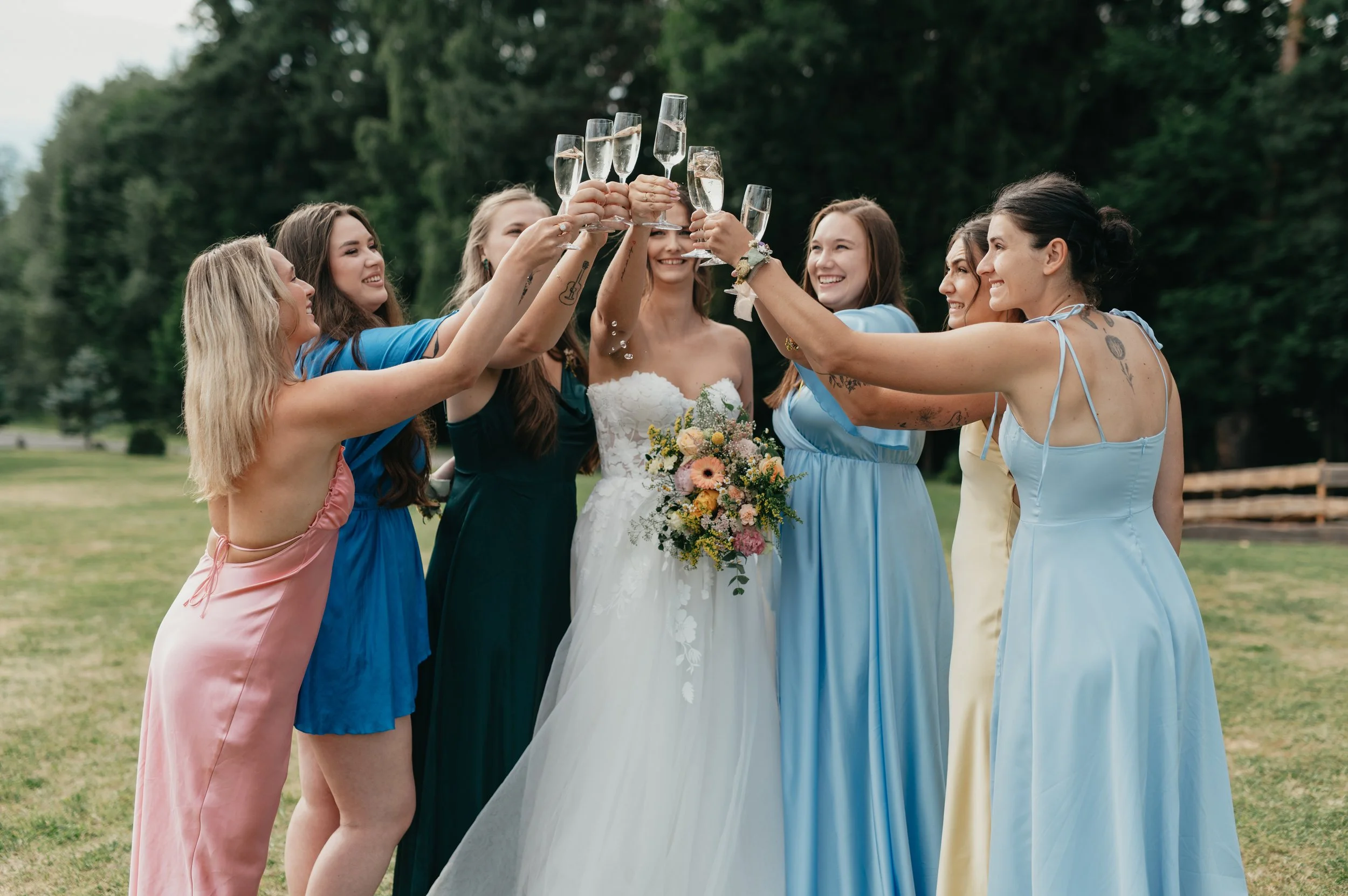 Group of women, including a bride in a white wedding dress holding a bouquet, raising champagne glasses in a toast outdoors in a lush green park, celebrating at a wedding.