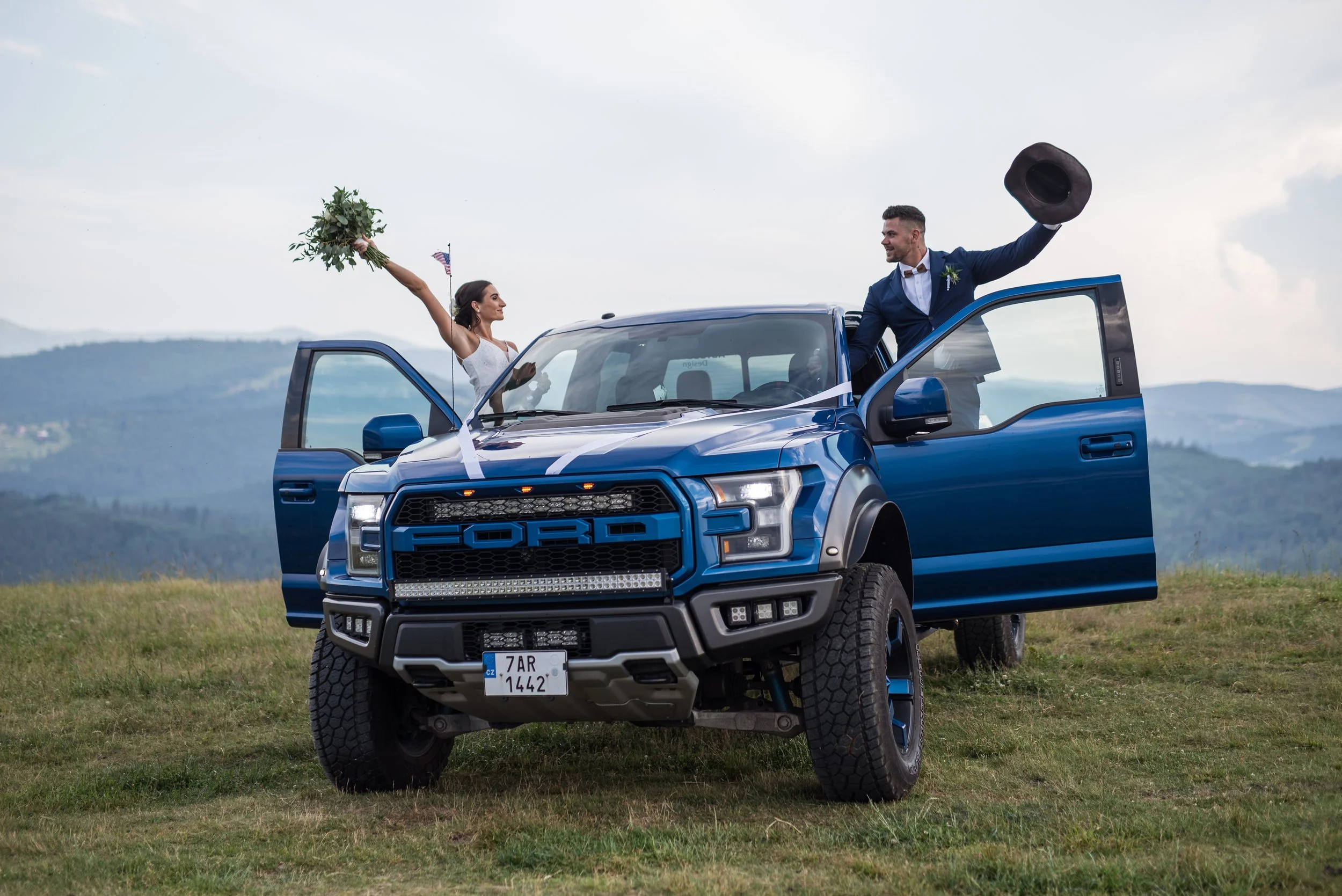 Bride and groom celebrating on a blue Ford truck in a scenic landscape.