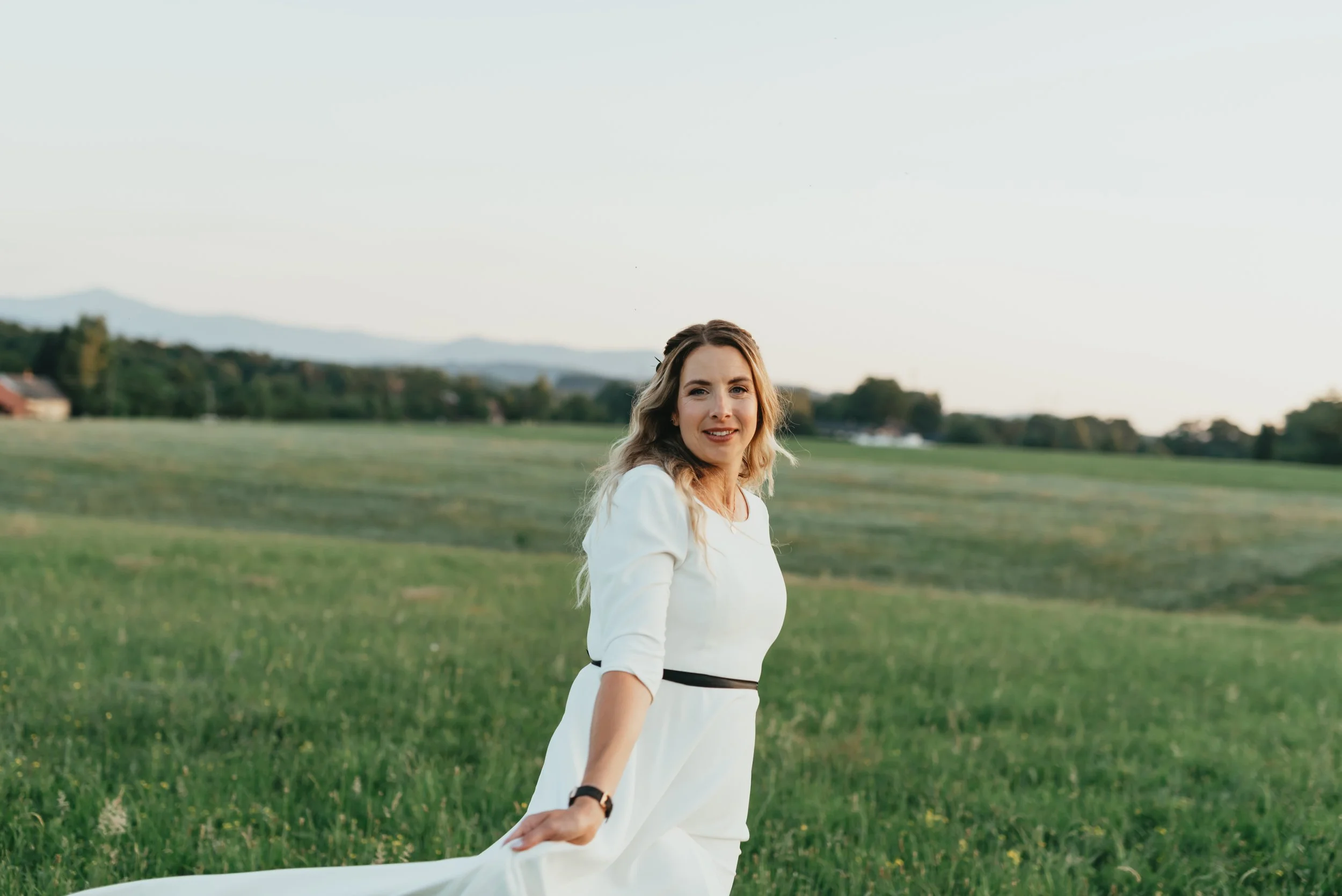 Woman in a white dress in a green field with mountains in the background.