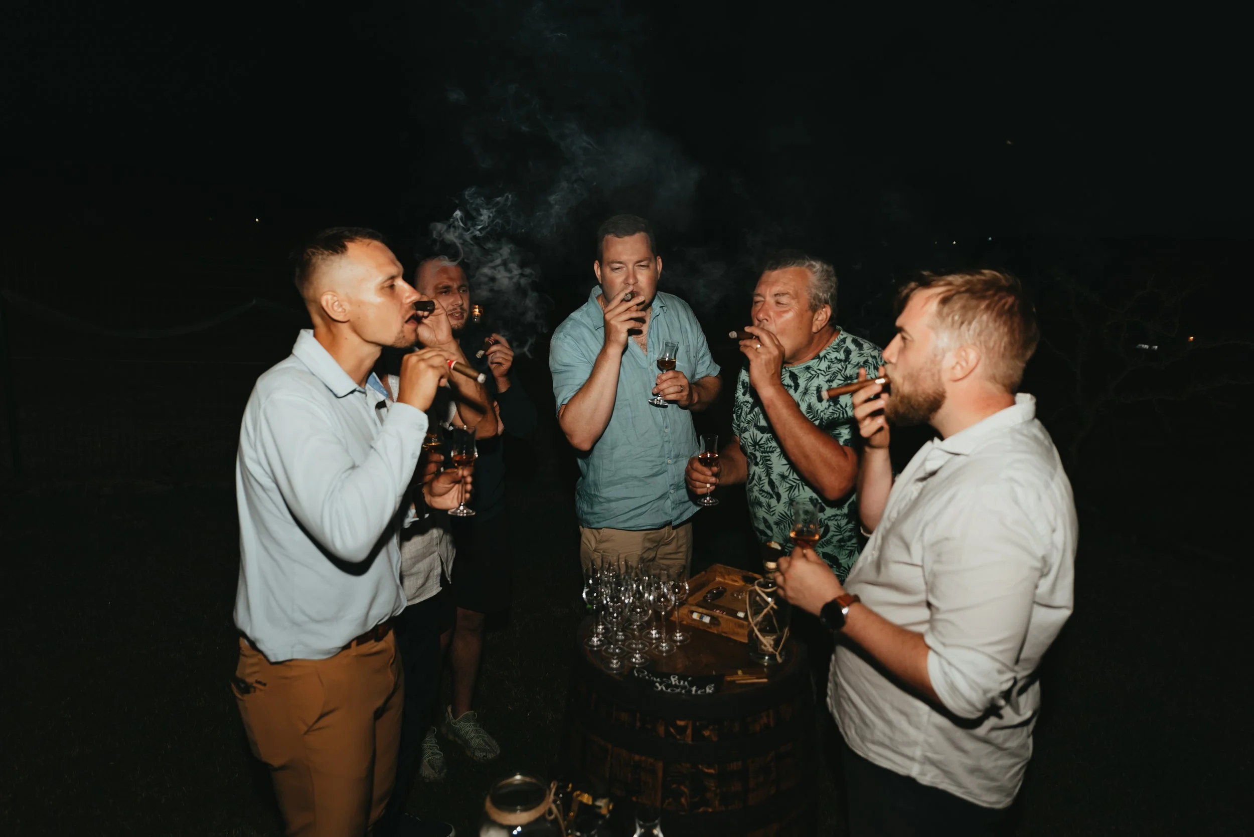 Group of men smoking cigars and holding drinks outdoors at night.