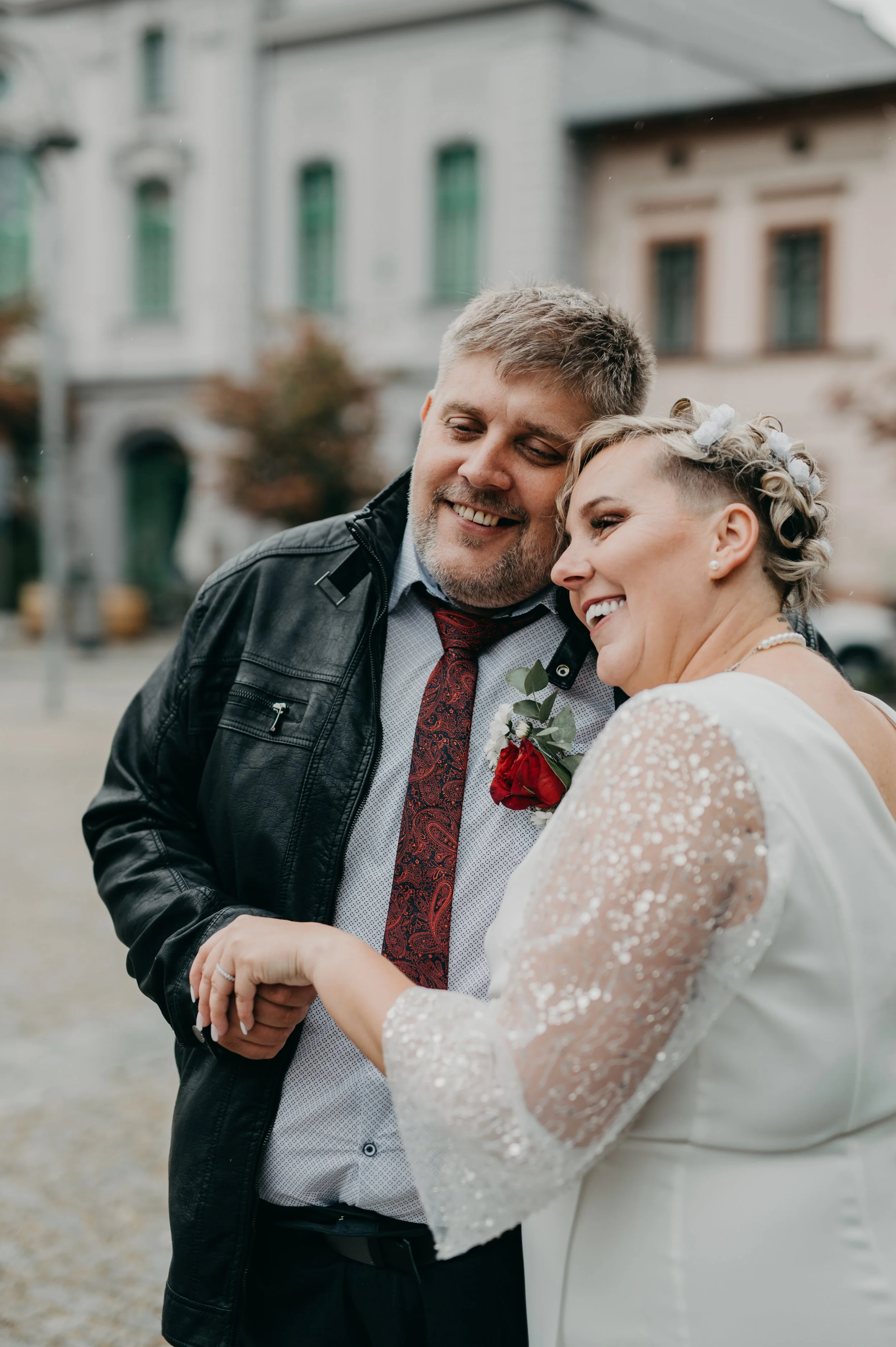 A happy couple with the woman in a white dress with sparkly details and the man in a black leather jacket, holding hands and smiling outdoors with buildings in the background.