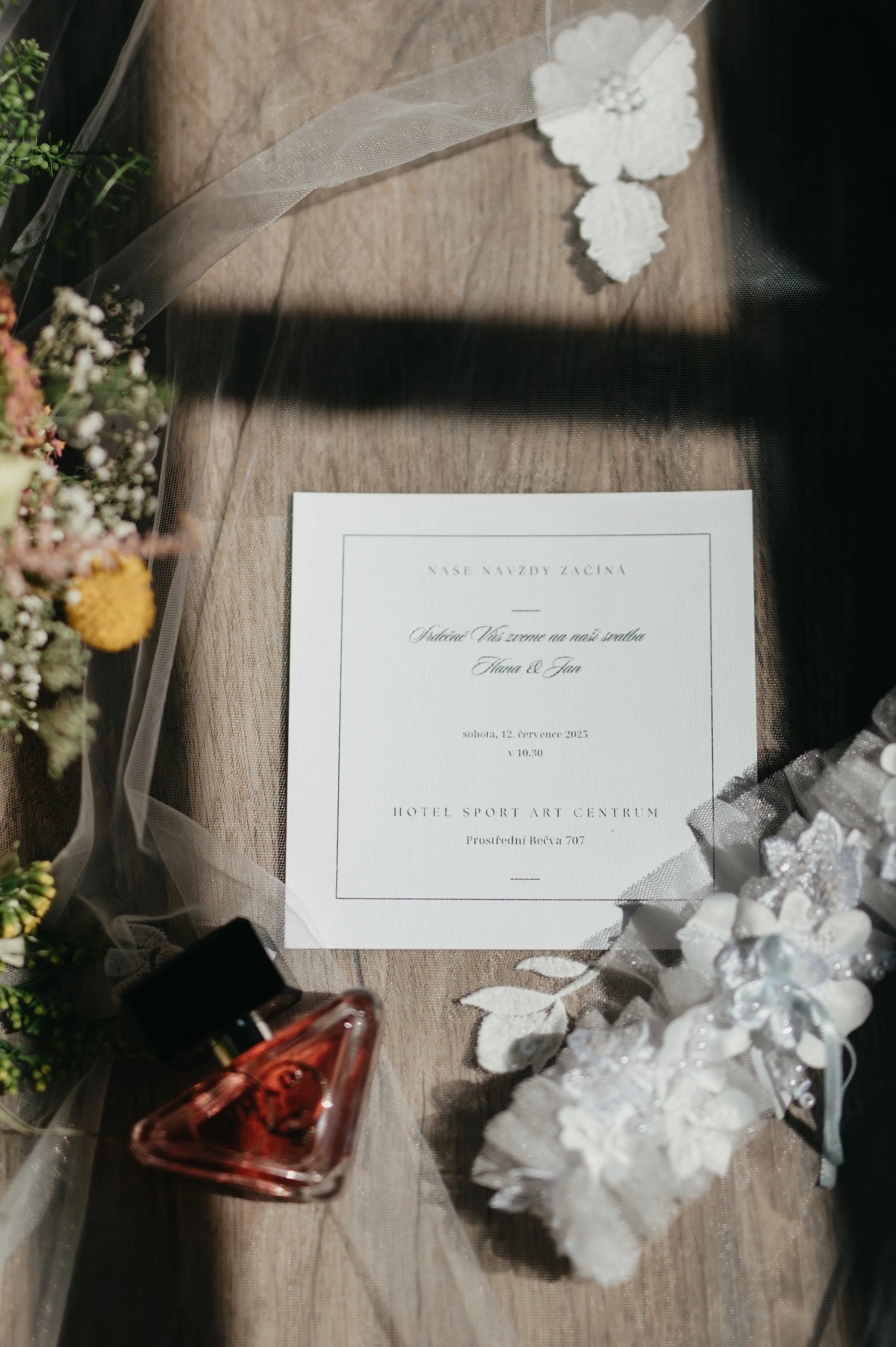 Wedding invitation on a wooden surface with a veil, a glass perfume bottle, and decorative white lace and floral elements.