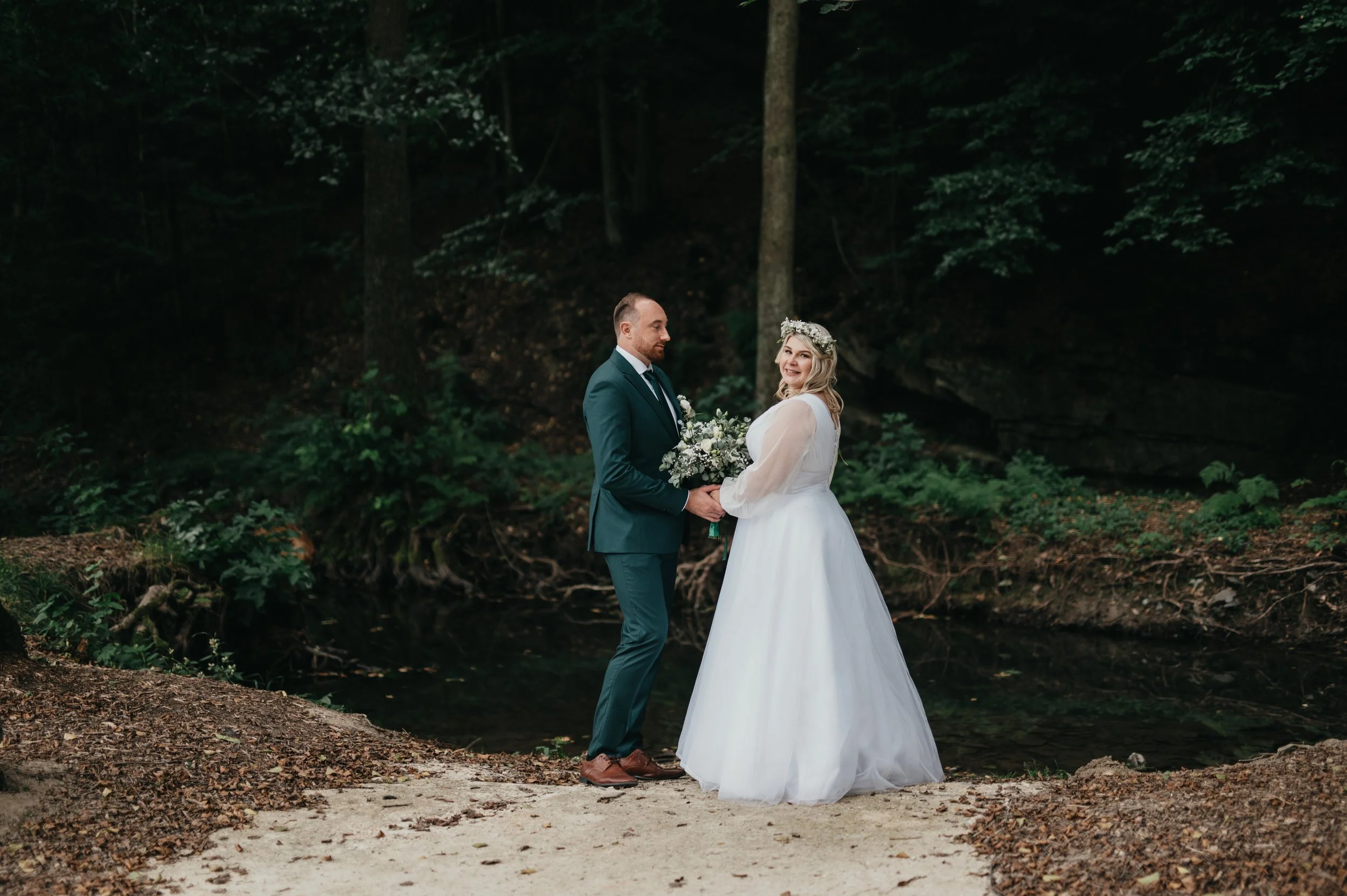 A bride and groom standing together outdoors in a forested area, holding a bouquet of flowers, during their wedding.