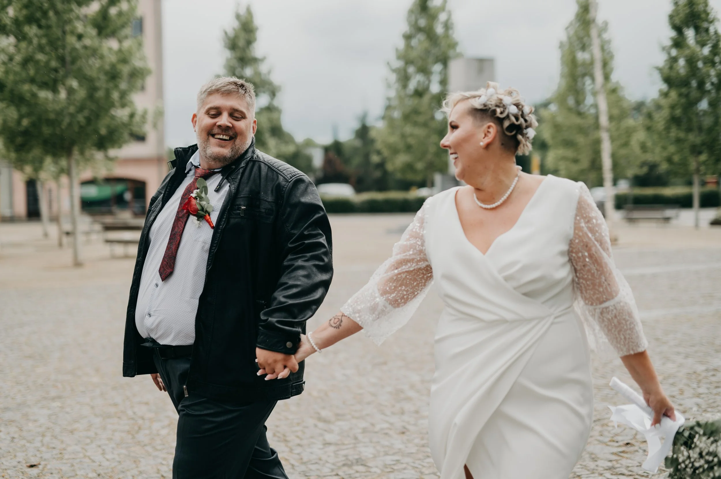 A smiling couple dressed in wedding attire, holding hands and walking outdoors in a paved area with trees and buildings in the background.