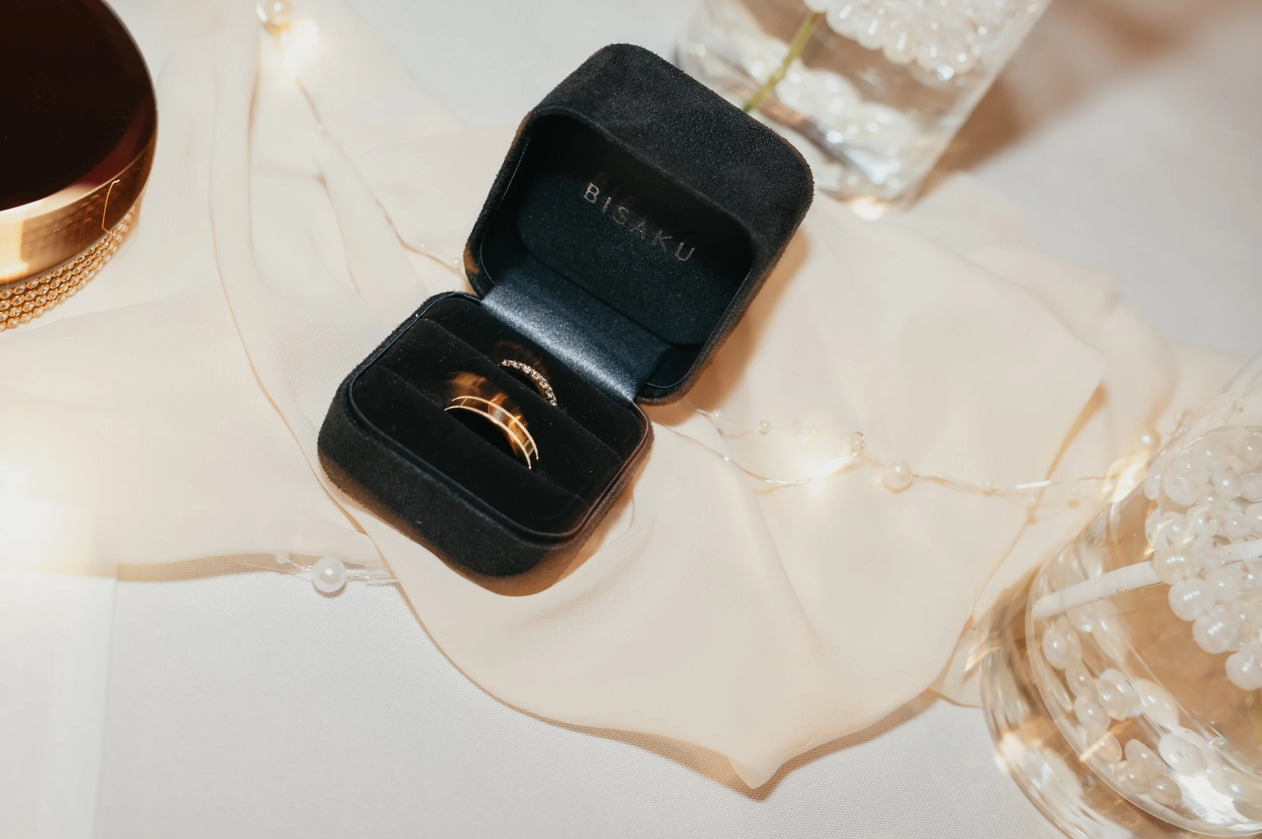 Černá sametová krabička na prsteny se zlatým prstenem uvnitř, umístěná na bílém stole s ozdobnými světýlky, sklenicí vody s ledem a nádobou s bílými korálky.