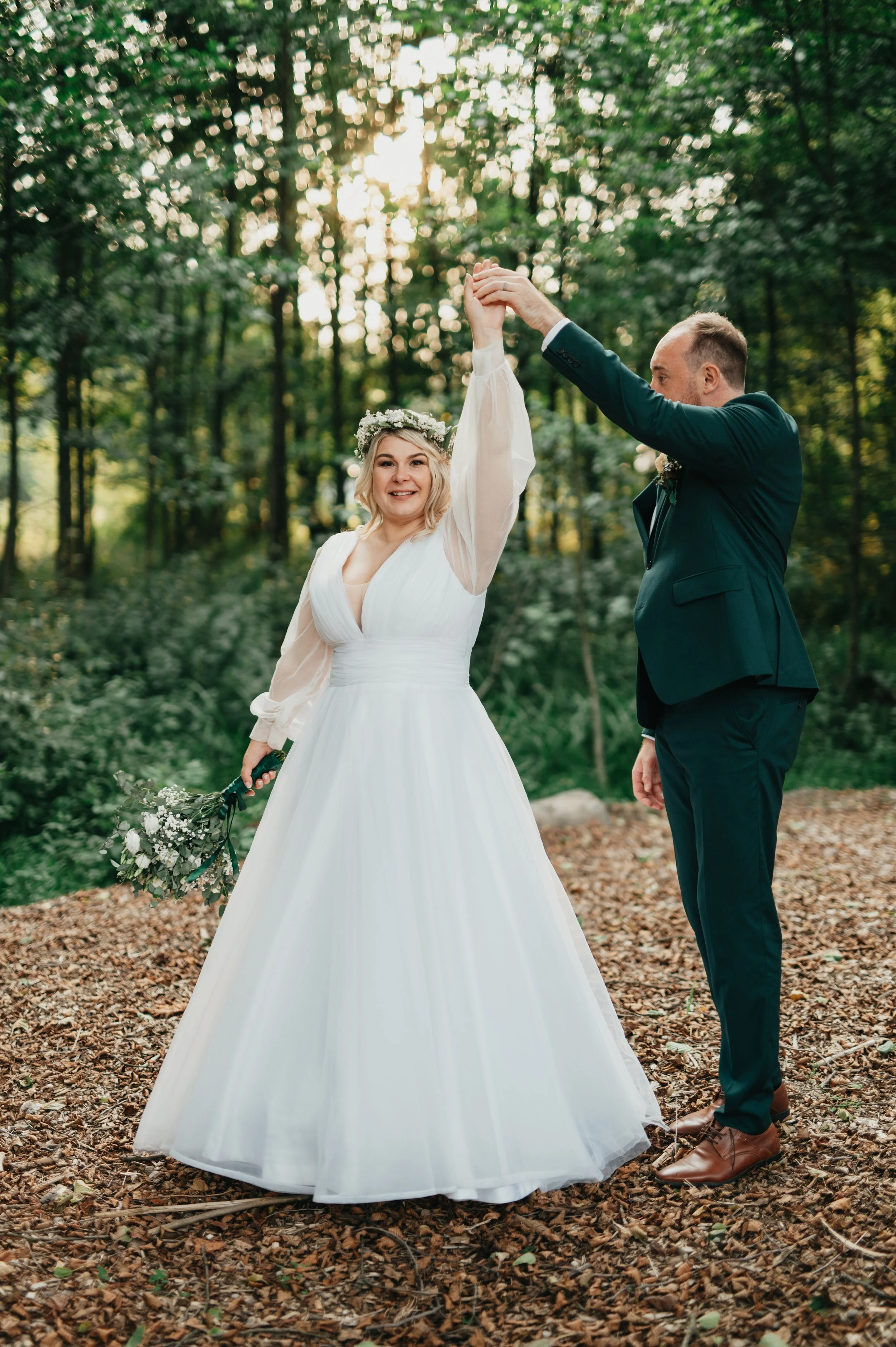 A bride and groom dancing in a forest during their wedding, with the bride wearing a white wedding dress and flower crown and the groom in a dark suit.
