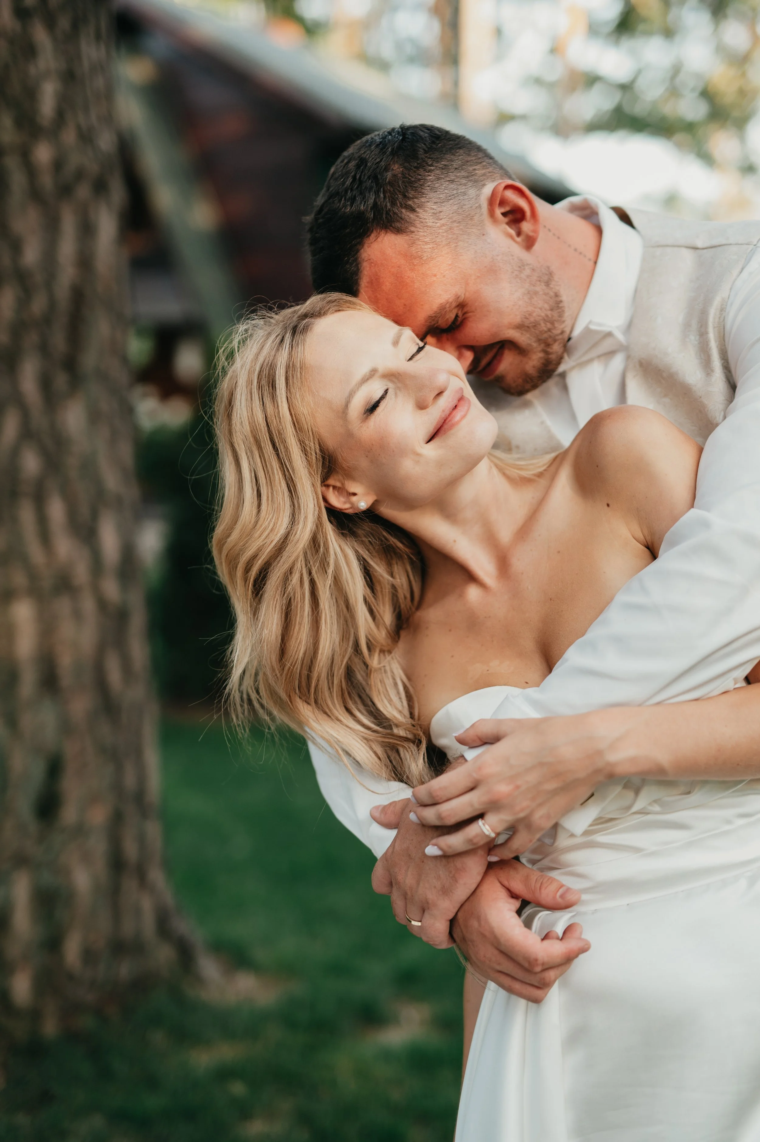A couple shares a romantic embrace outdoors, with the man on top and the woman smiling with her eyes closed. They are dressed in wedding attire, standing near a tree with blurred background of greenery and a house.