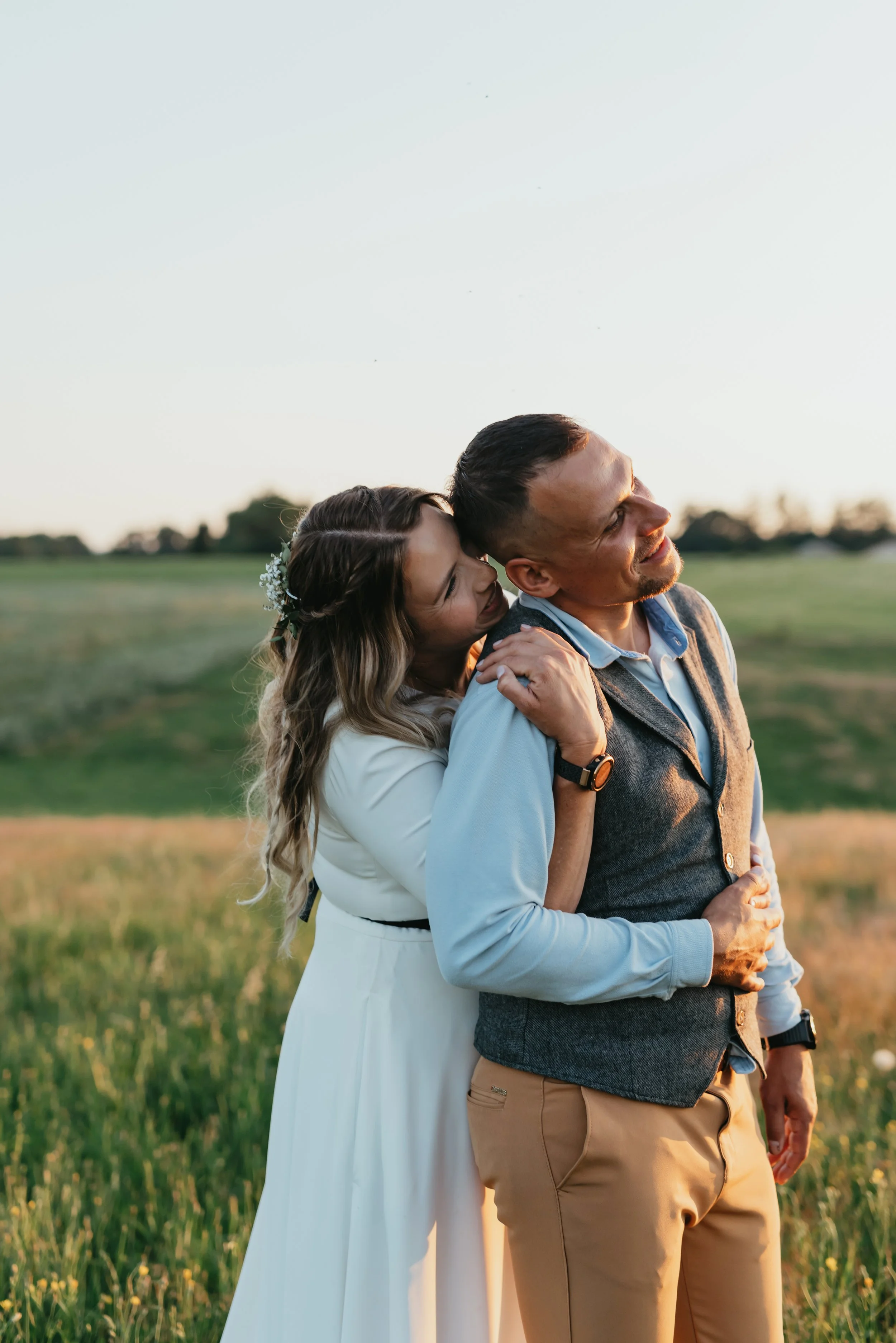 A couple embracing in a grassy field during sunset, with the woman hugging the man from behind. She is wearing a white dress and has flowers in her hair. He is in a blue shirt and gray vest, both smiling.