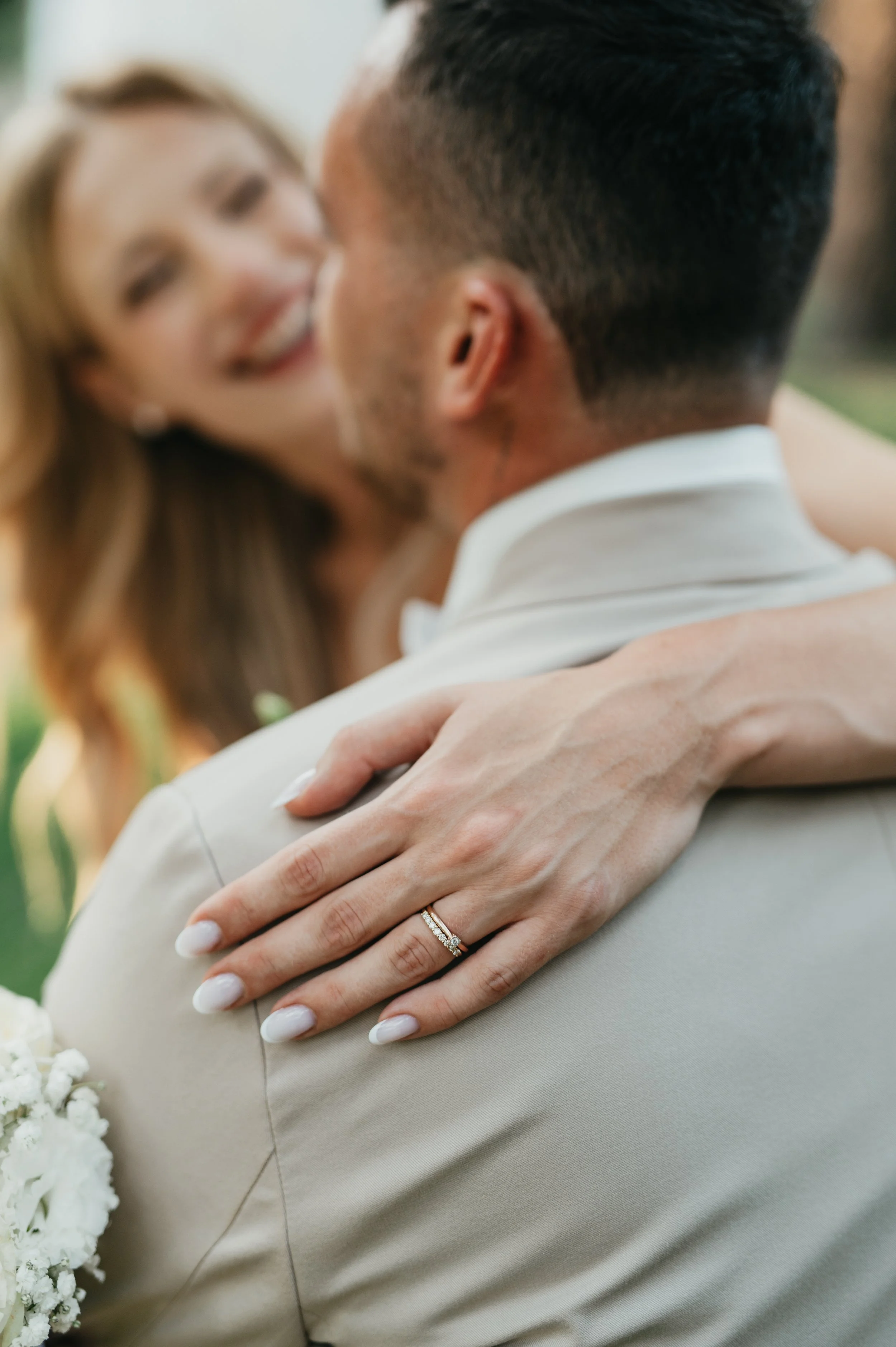 A woman with a wedding ring on her finger embraces a man, with a blurred woman in the background, at an outdoor wedding or celebration.