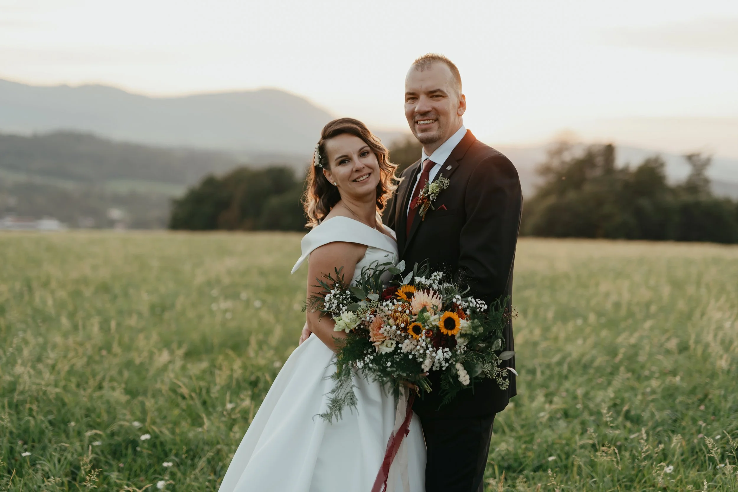 Bride and groom in a field holding a bouquet, smiling at sunset with mountains in the background.