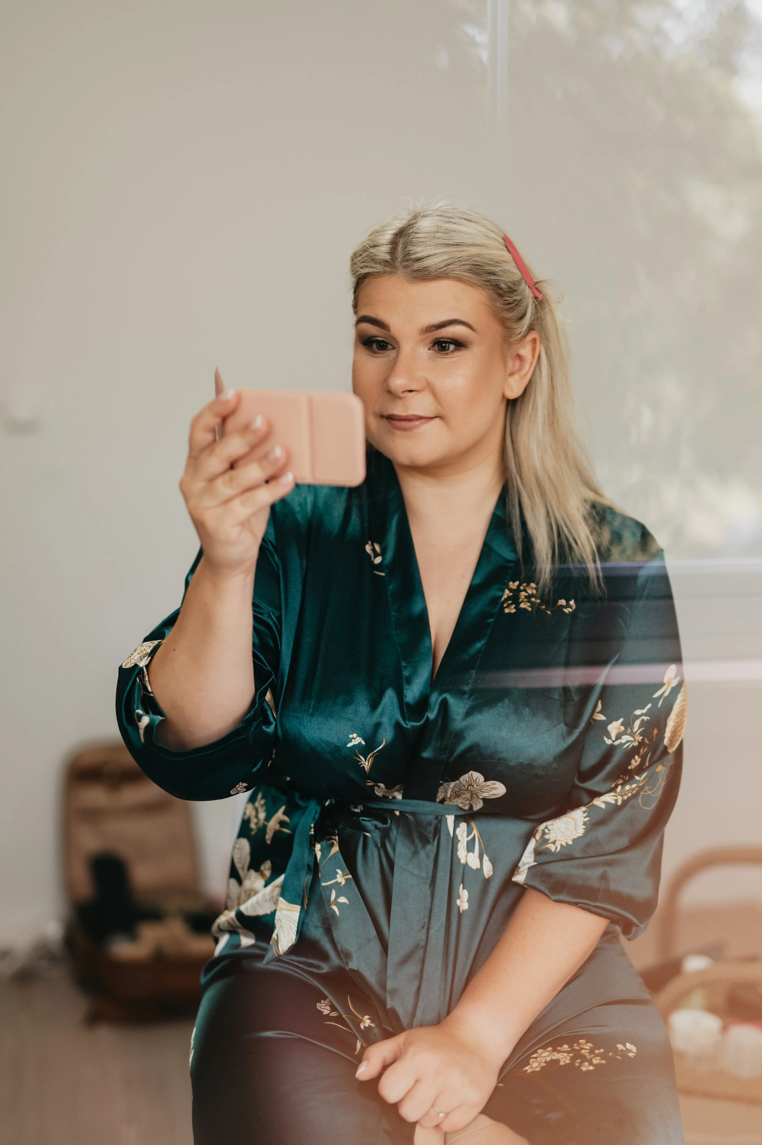 A woman with blonde hair wearing a floral satin robe is sitting and looking at her phone, appearing to take a selfie or video.