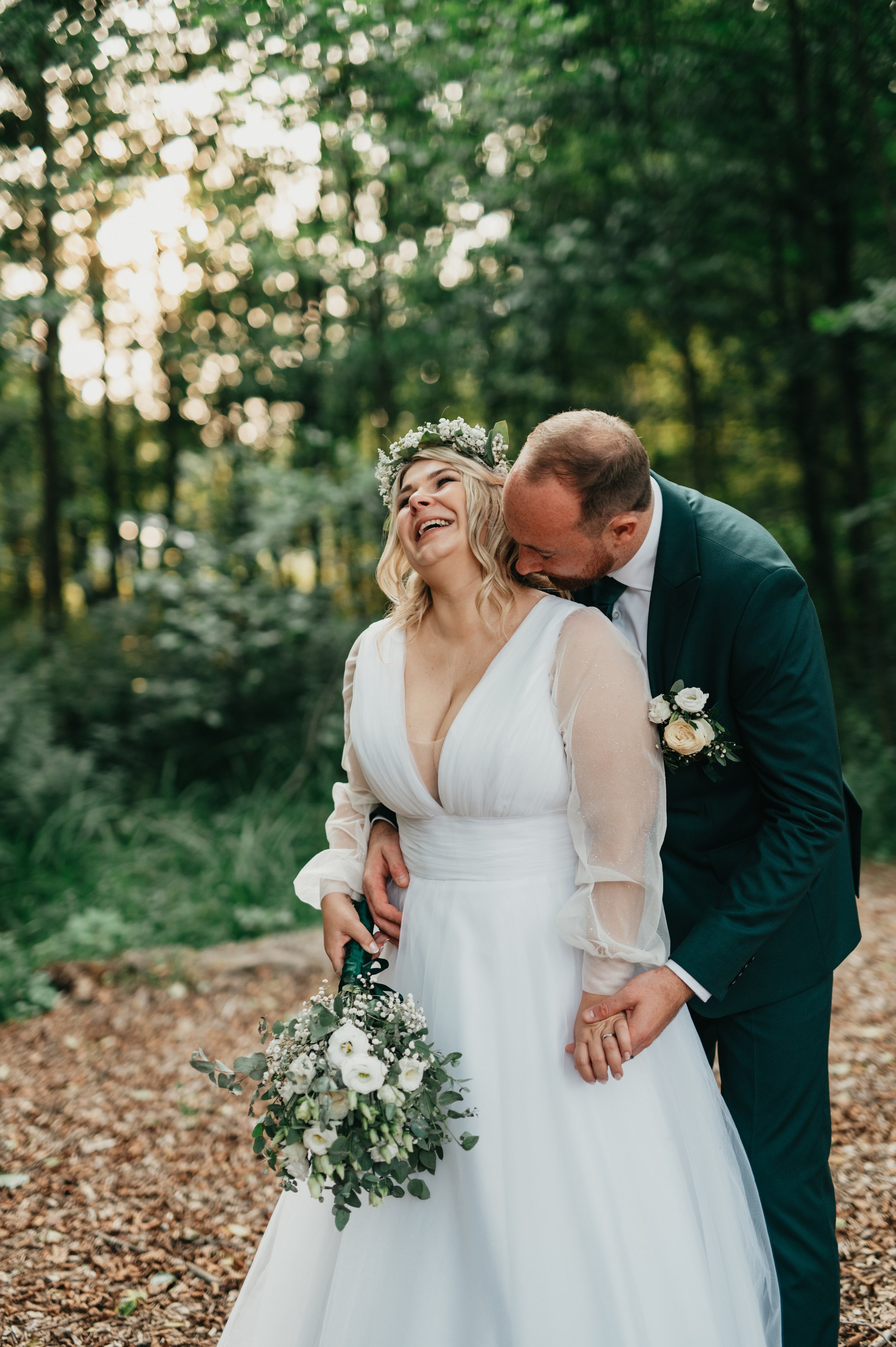 A happy bride and groom sharing a joyful moment outdoors in a wooded area during their wedding. The bride is smiling with her eyes closed, wearing a white dress with sheer sleeves and a floral crown, holding a bouquet of white flowers and greenery. T