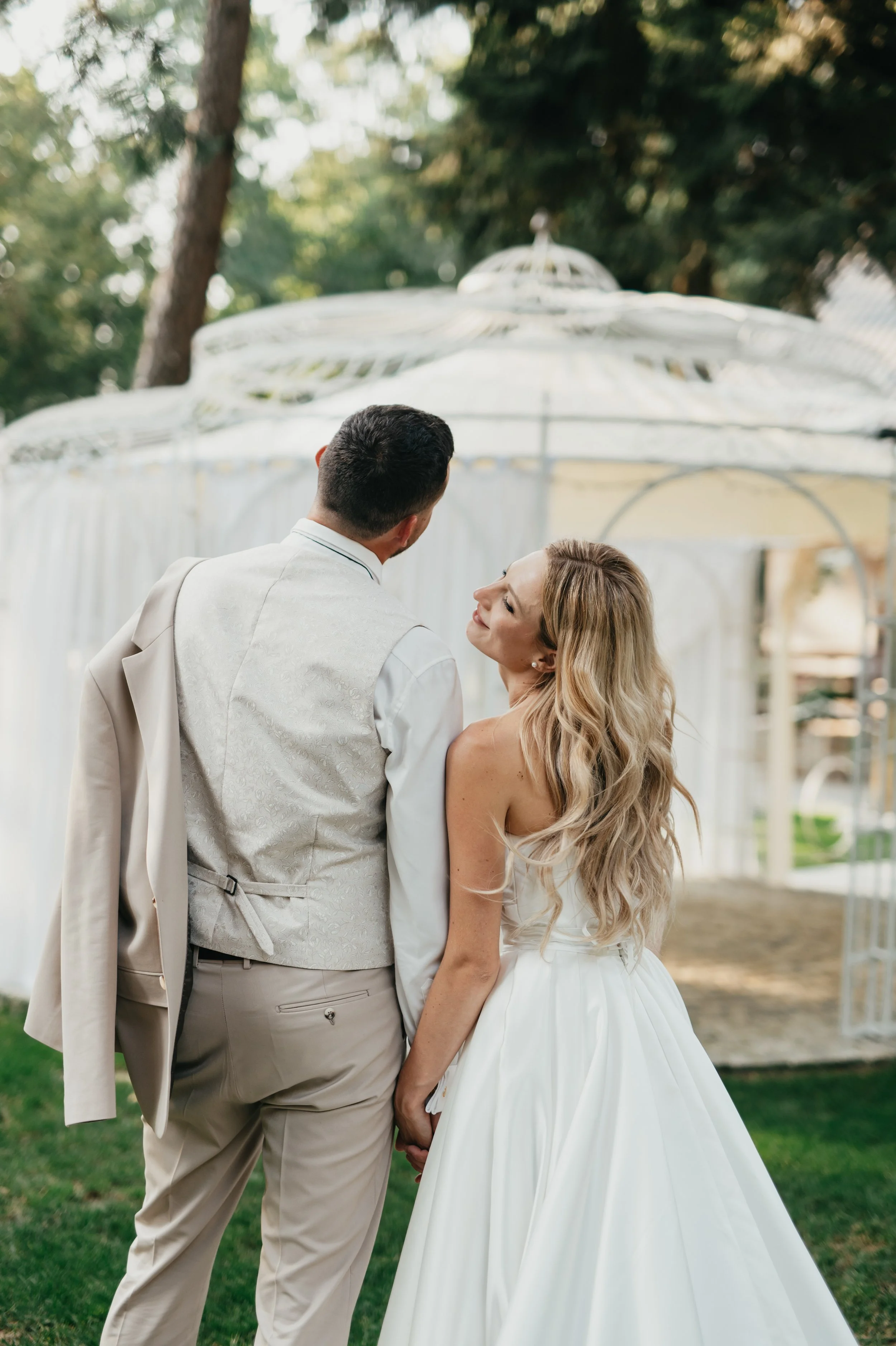 Bride and groom holding hands, standing outdoors in front of a white gazebo.