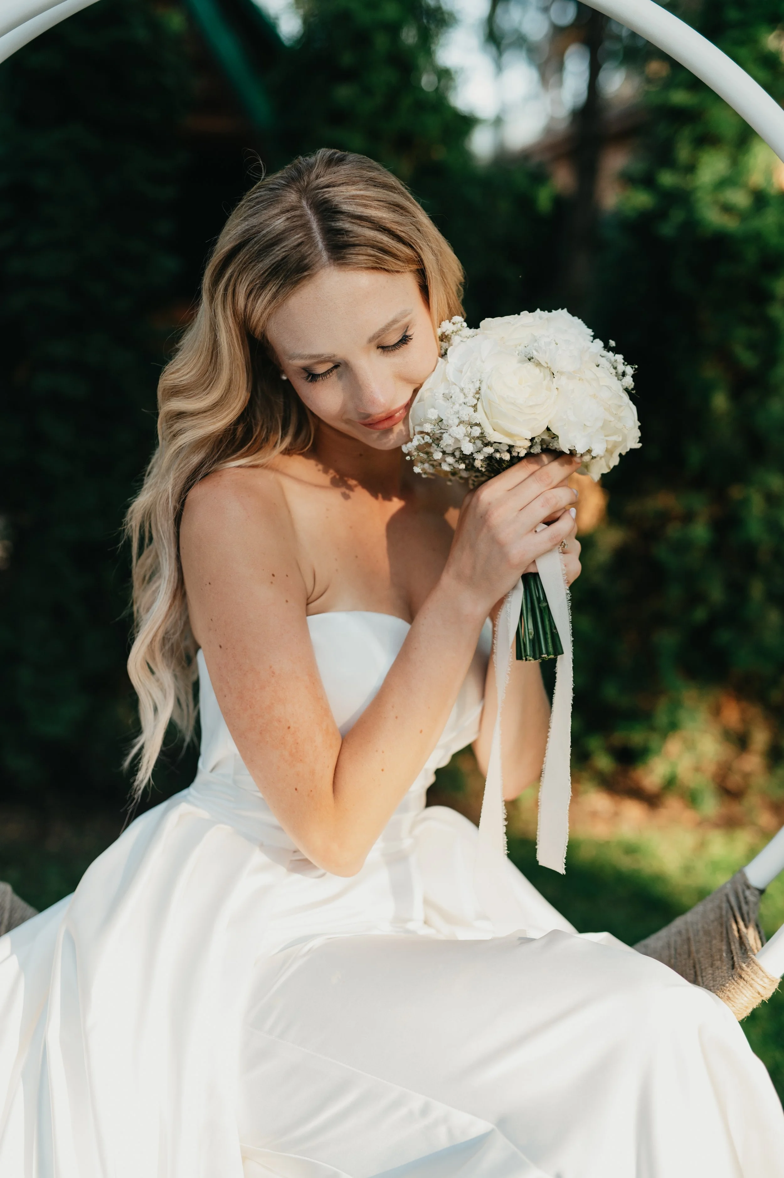 A bride in a strapless white wedding gown holding a bouquet of white roses and baby's breath, sitting outdoors with greenery in the background.