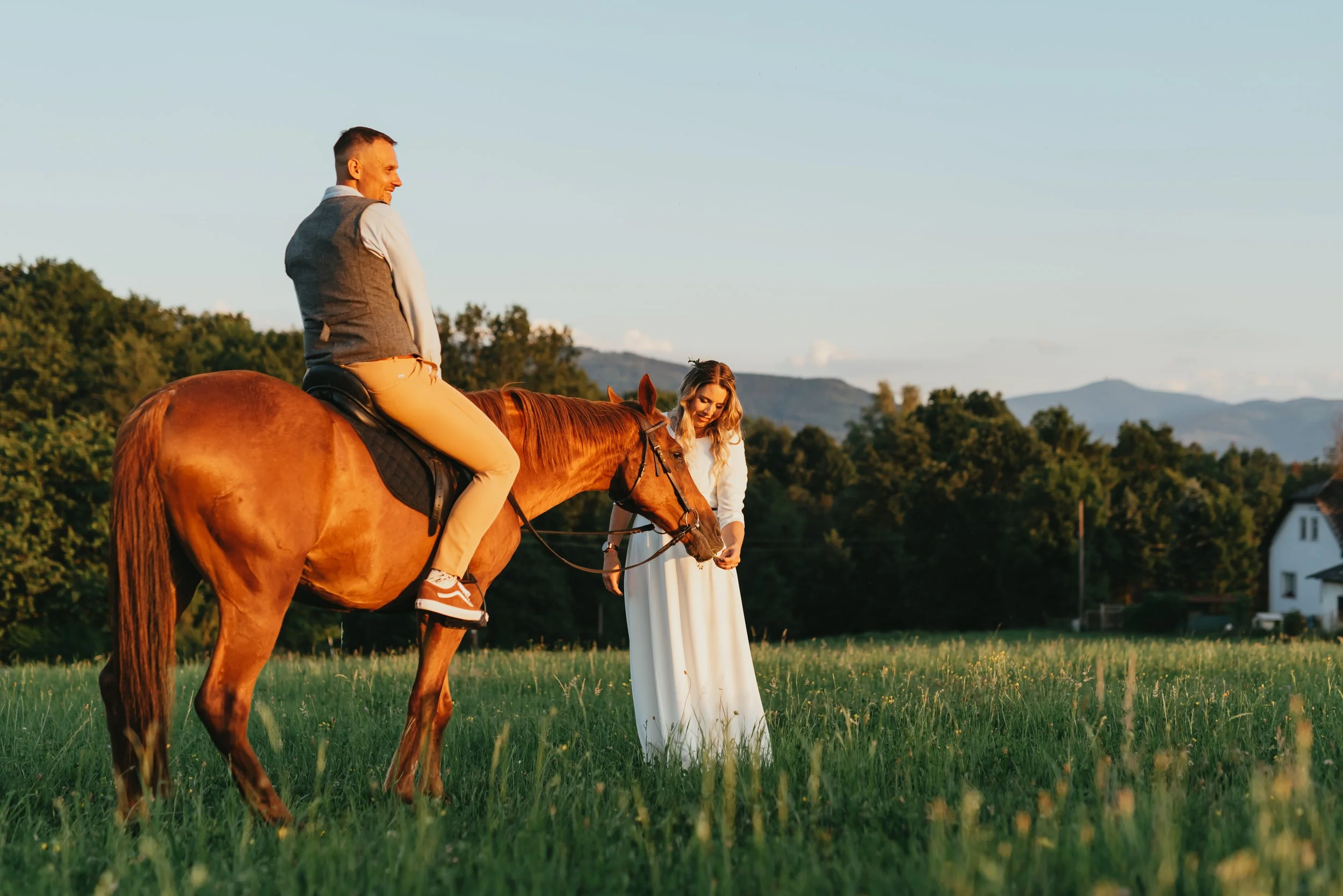 Man sitting on a horse and woman in a white dress standing in a grassy field with mountains in the background.