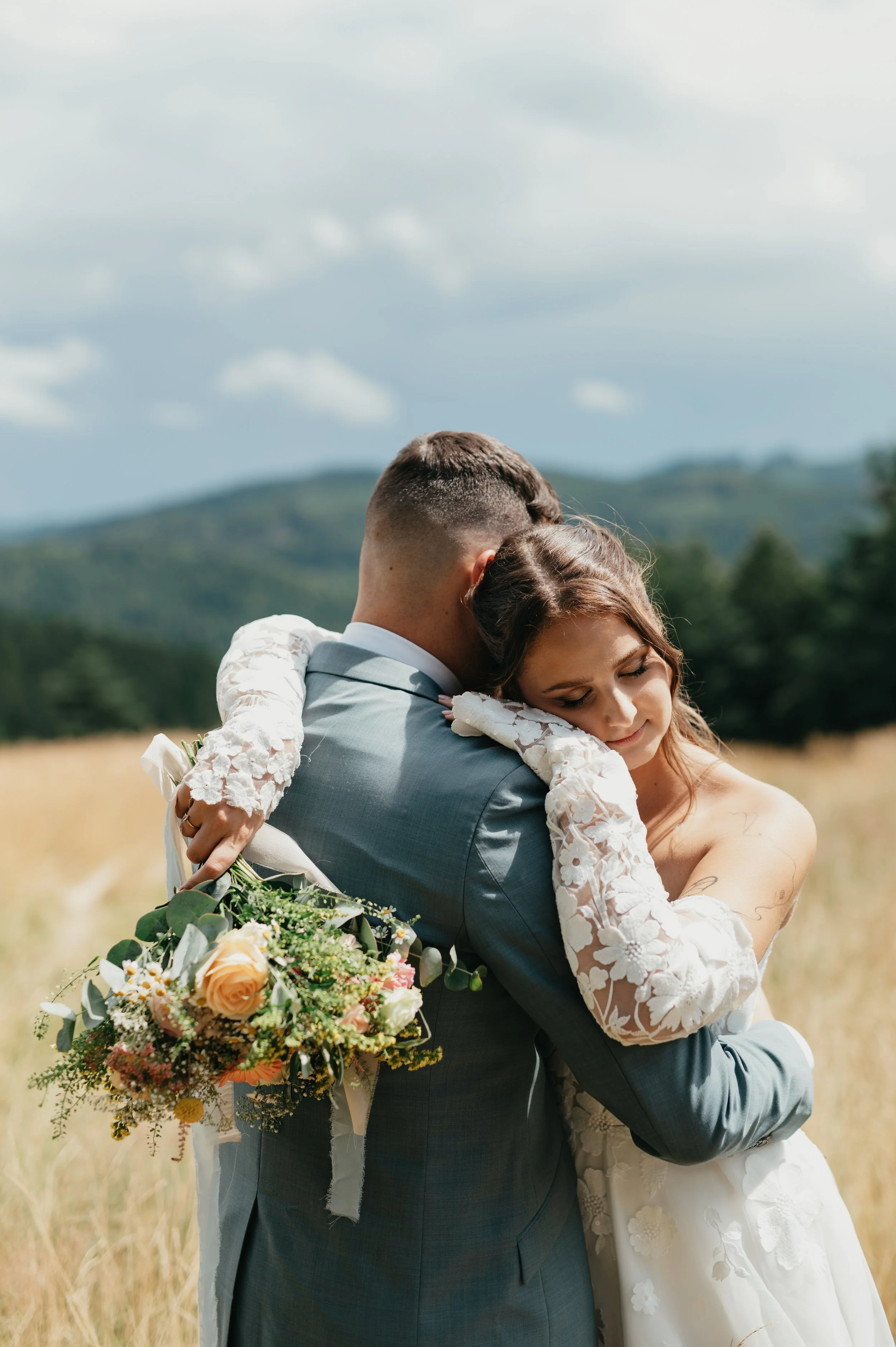 A bride and groom embrace outdoors in a field with mountains in the background, the bride holding a bouquet of flowers.