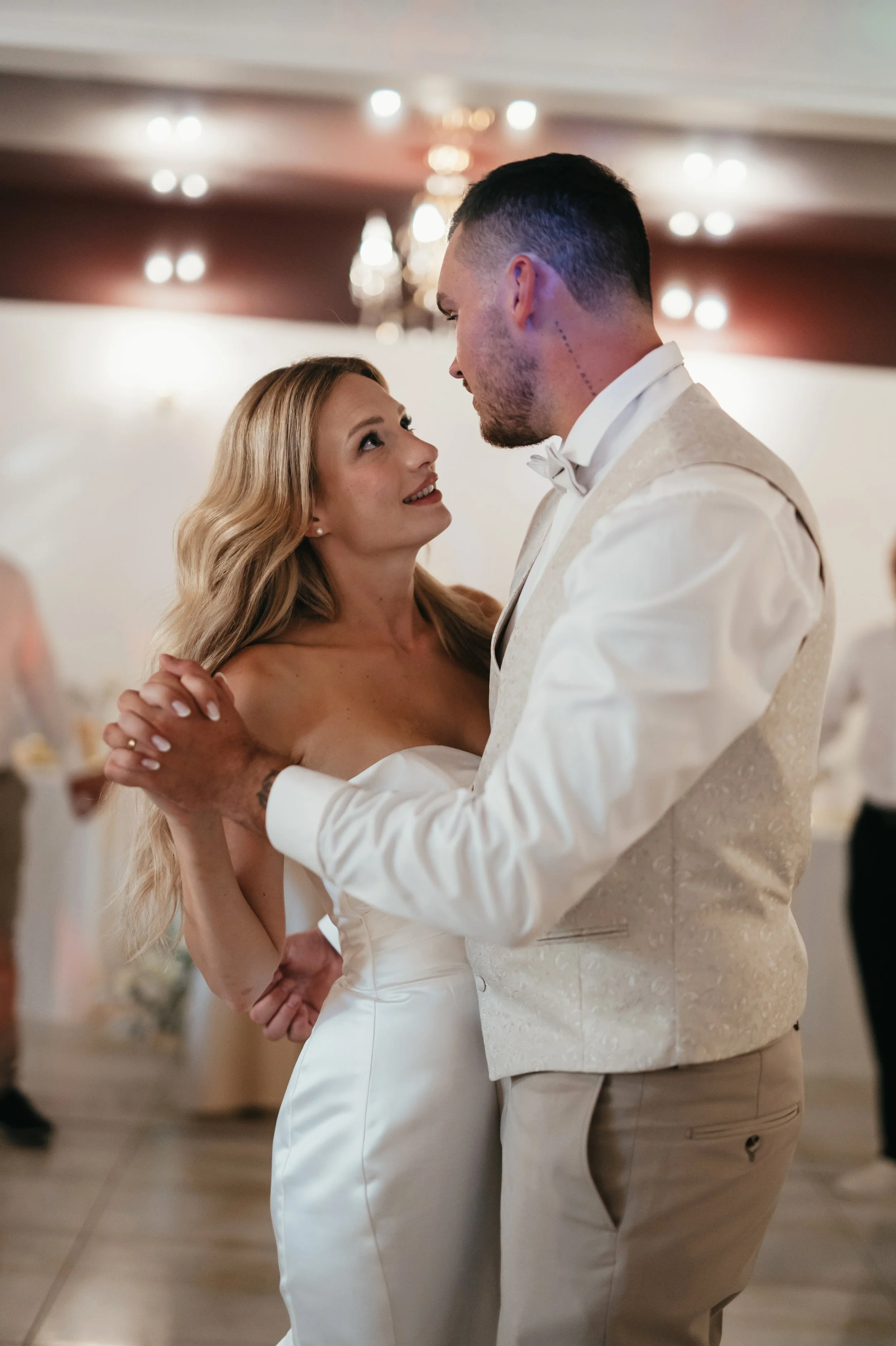 A bride and groom share a dance at their wedding reception indoors, with warm lighting and blurred background guests.