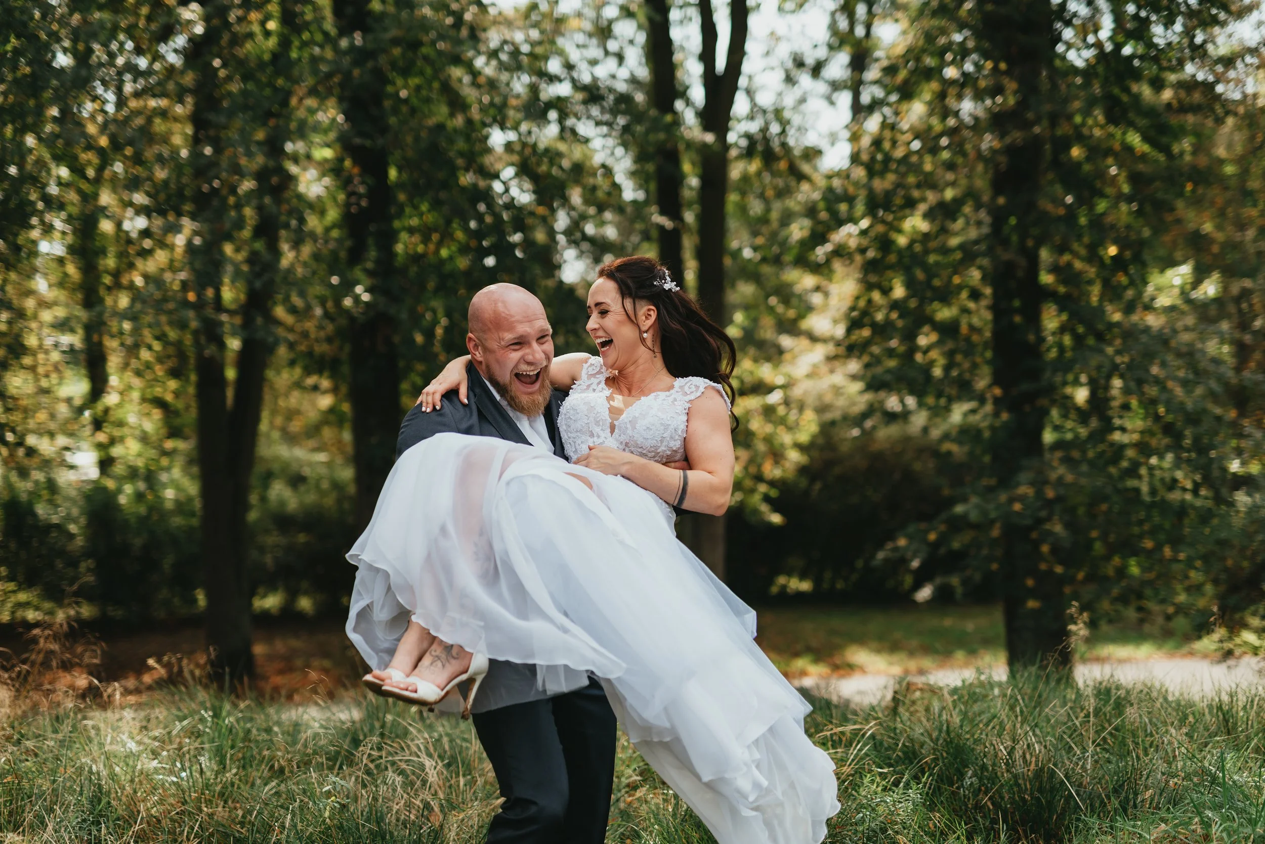 Smiling groom carrying bride in white dress in a forest setting.
