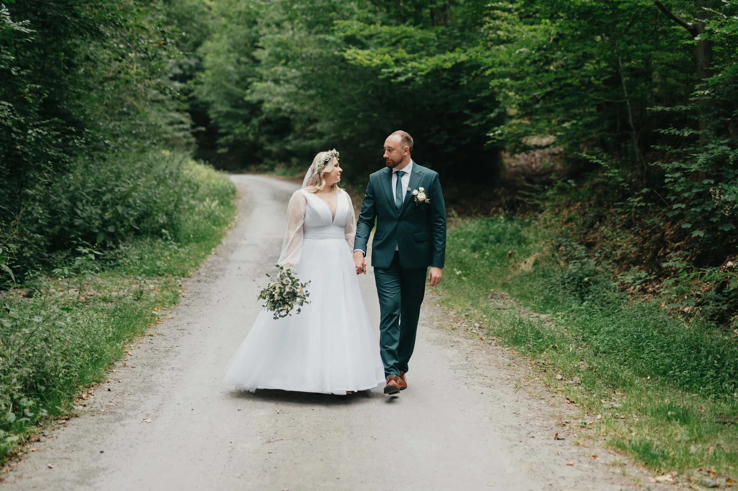 A bride and groom walking hand in hand along a gravel road through a green forest.