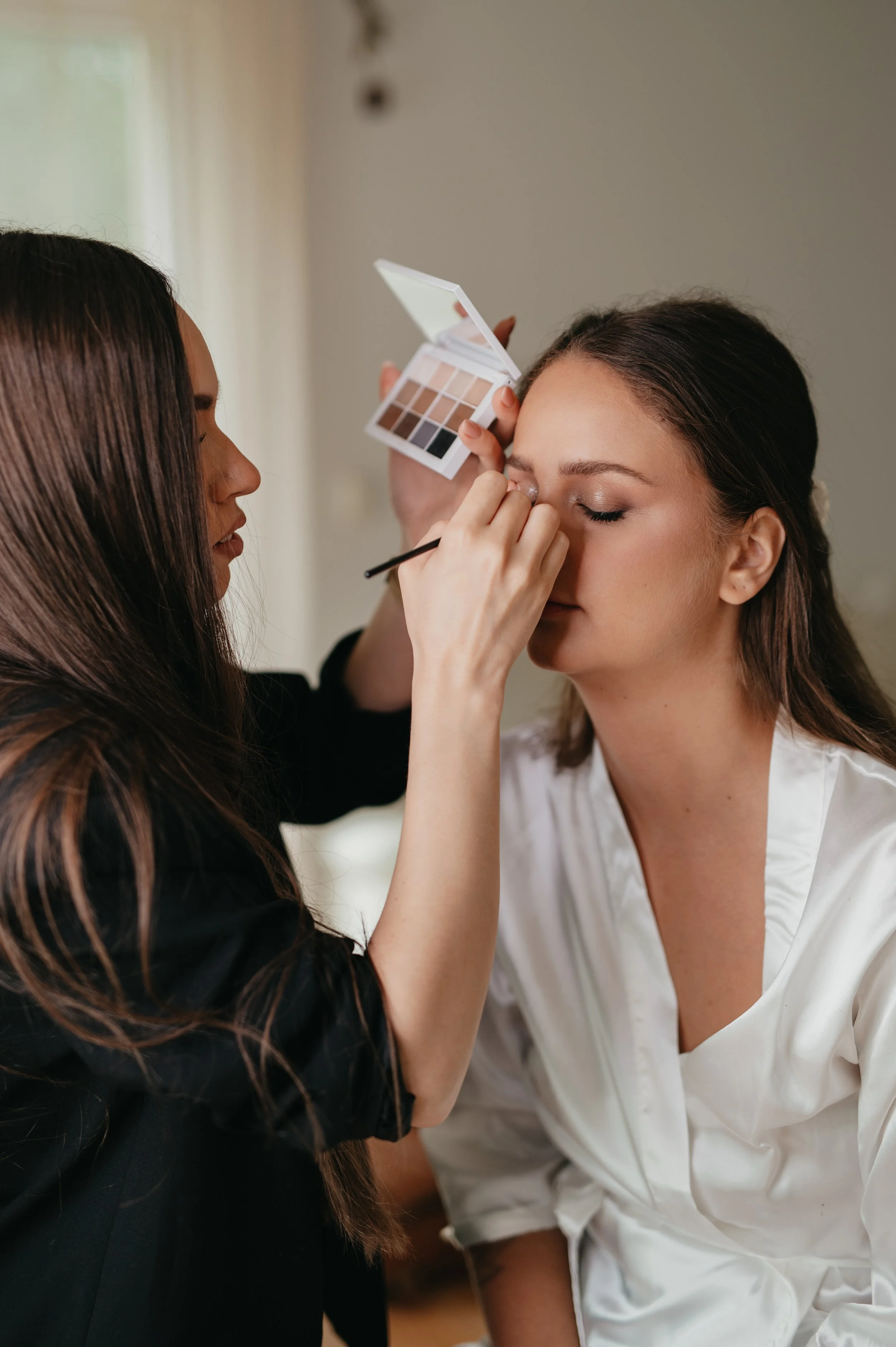 A woman applying makeup to another woman with closed eyes, holding an eyeshadow palette in her other hand.