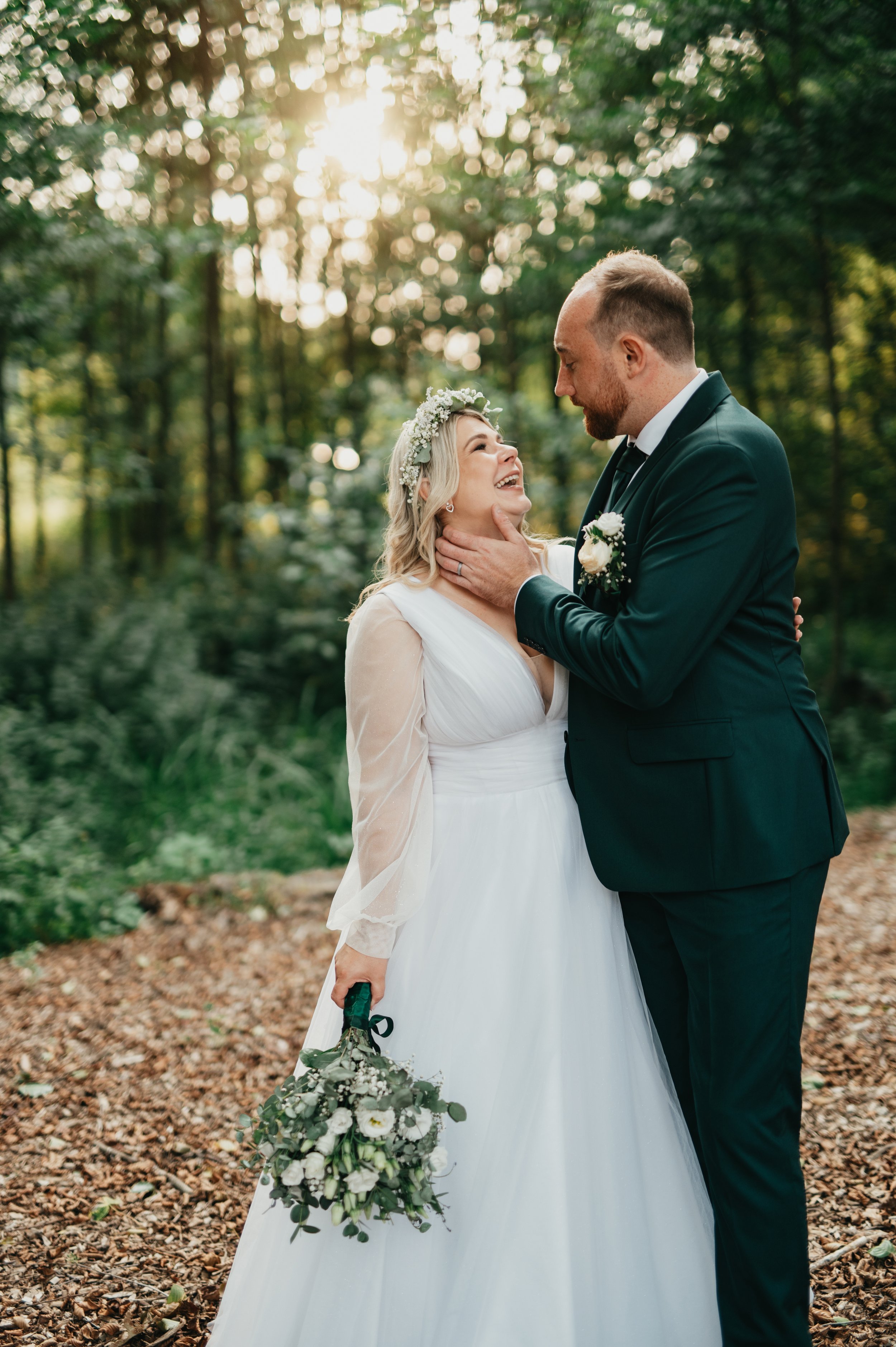 A newlywed couple in wedding attire standing in a forest during sunset, with the groom lifting the bride's chin and smiling at her.