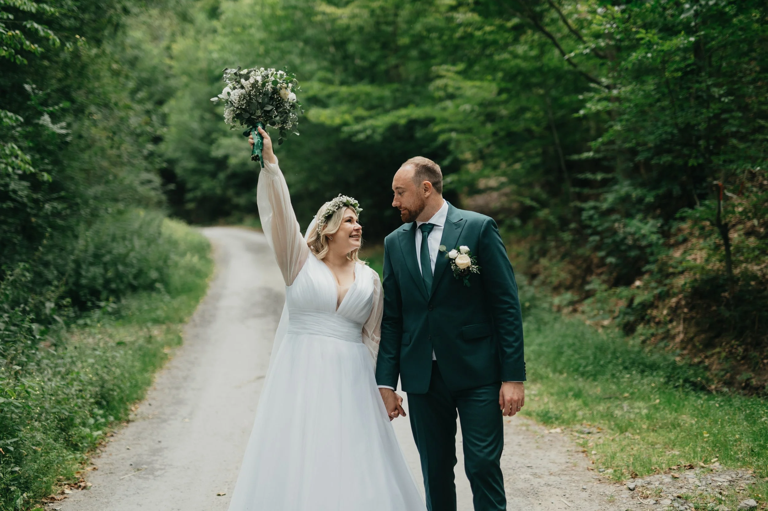 A newlywed couple holding hands on a dirt road surrounded by green trees in a forest, with the bride raising her bouquet in celebration.