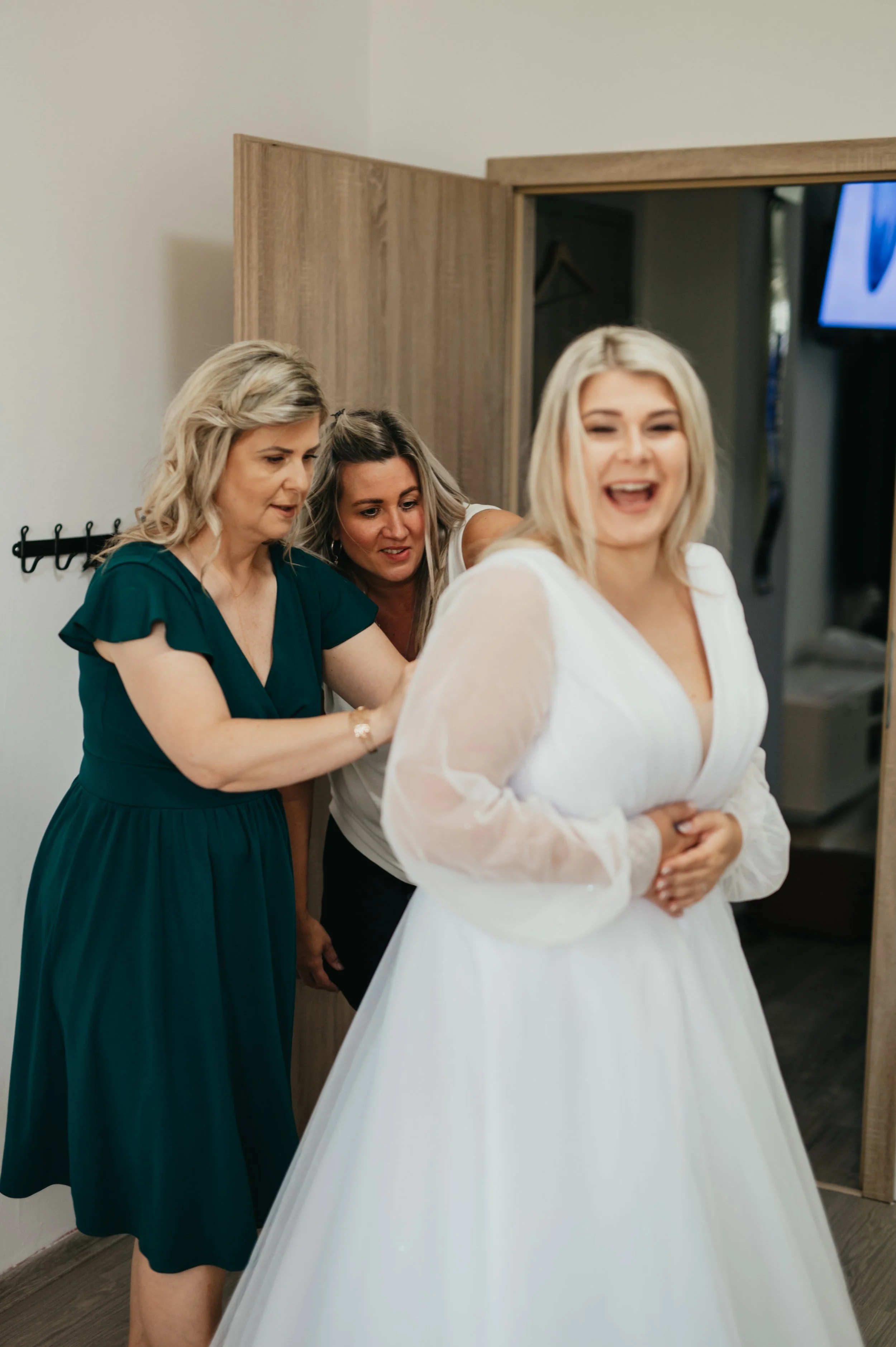 A bride in a white dress laughing as her bridesmaids help her get ready.
