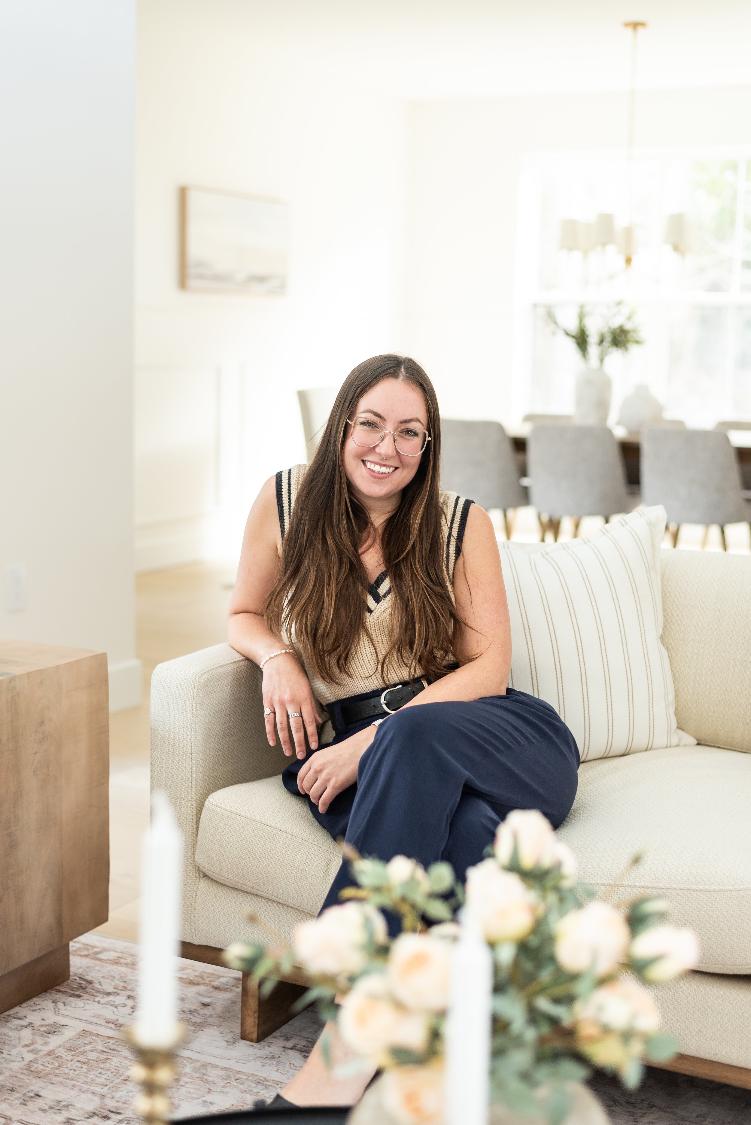 A stager a Charlotte Interiors. A young woman with glasses, long brown hair, and a white sleeveless top sitting on a bed in a bedroom with a beige wall, a framed landscape picture, a striped pillow, and a bedside table with a lamp.
