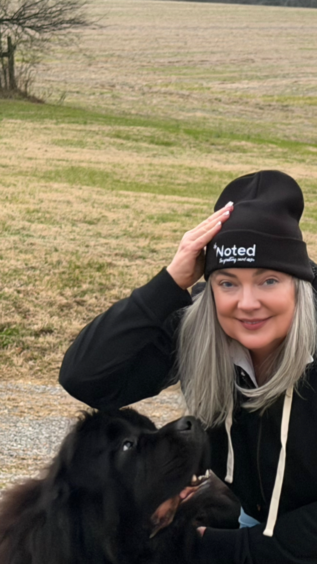 A woman with blonde hair smiling, wearing a black beanie and black jacket, petting a black dog outdoors with grass and fields in the background.