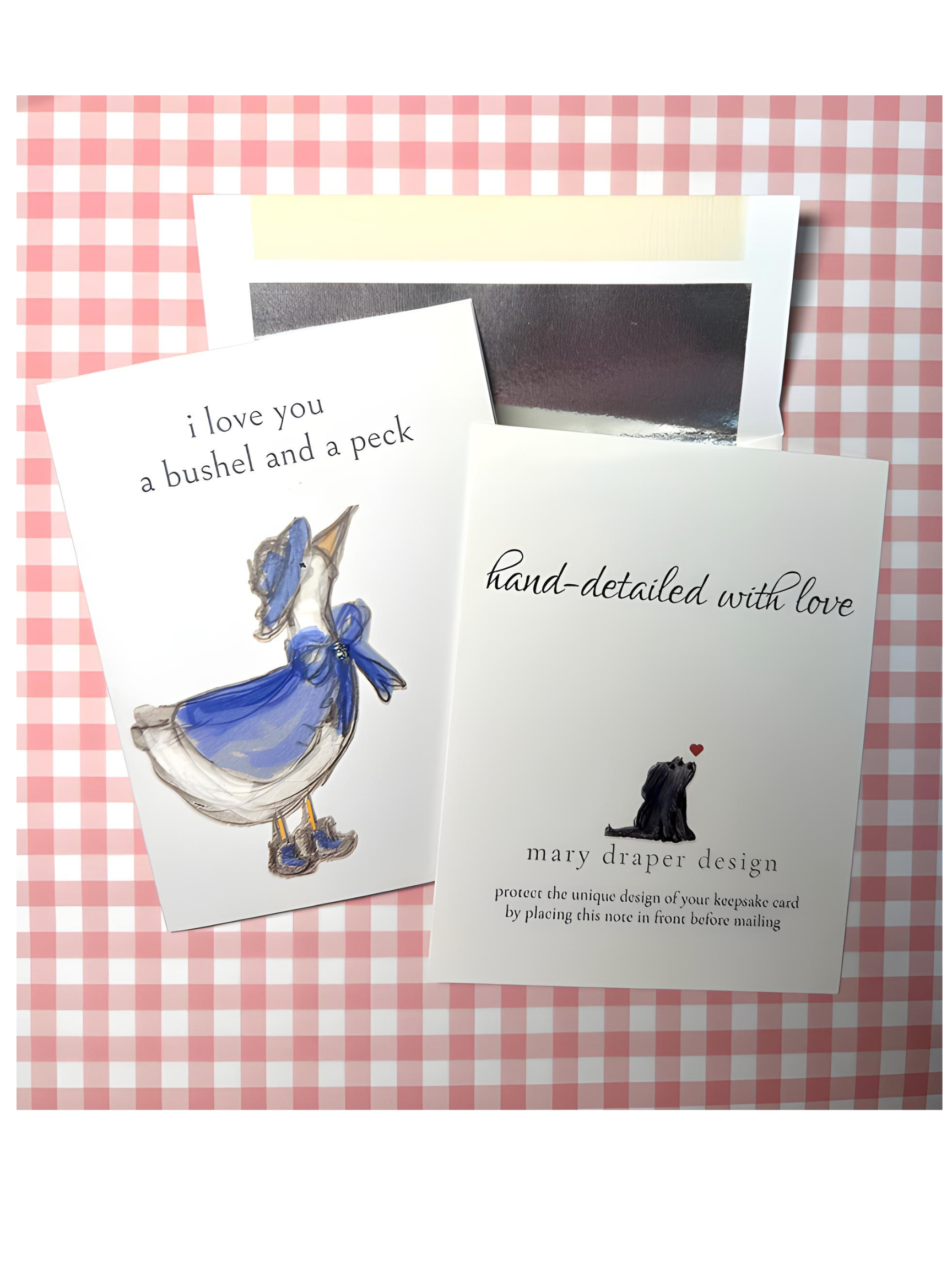 A greeting card printed on premium cardstock and hand-detailed from a watercolor illustration of a small duck in a blue dress tied with a bow and a hat. The text says 'i love you a bushel and a peck'. Silver-lined envelope.