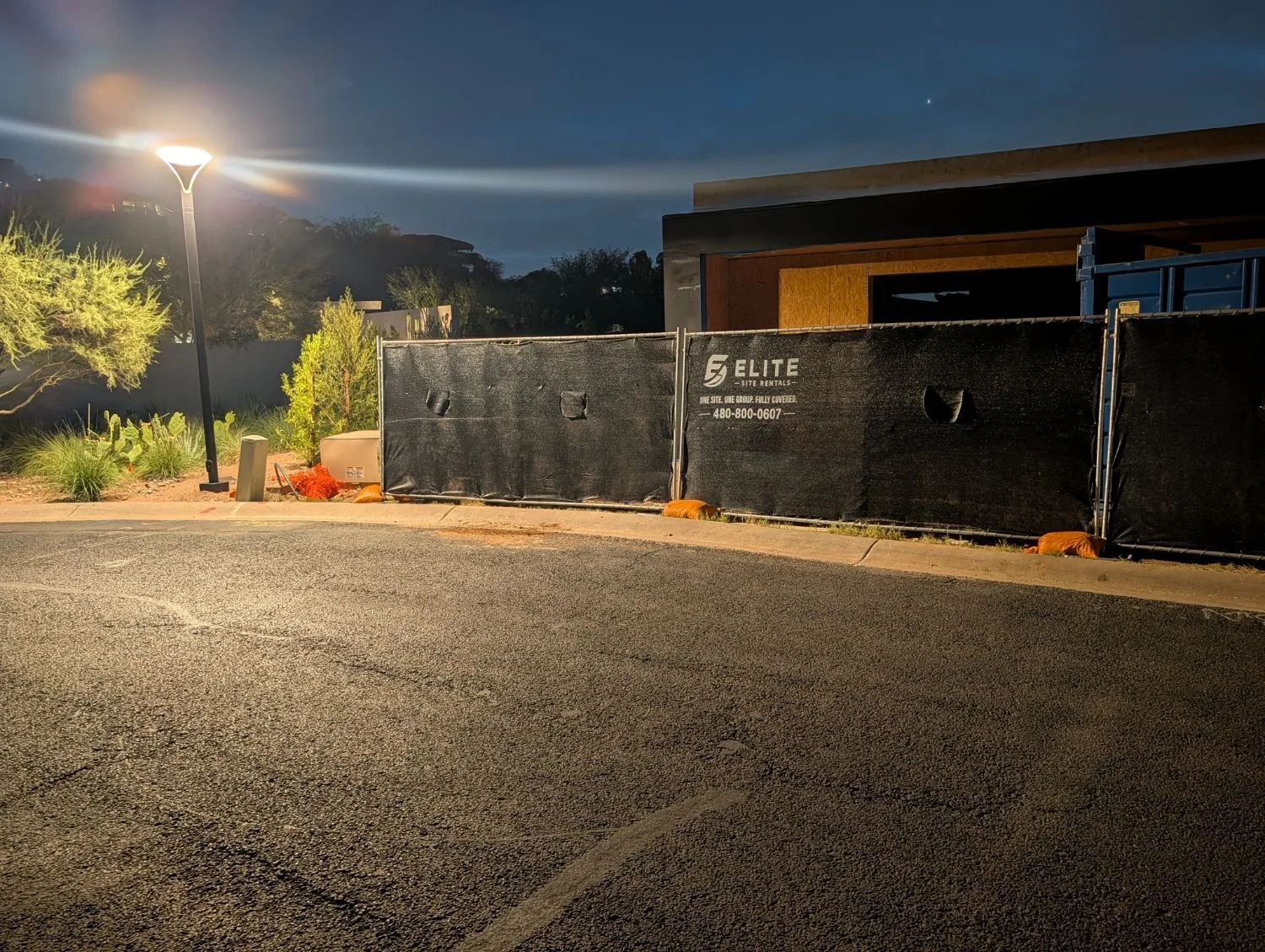 Nighttime view of a construction site with a black fencing barrier, construction materials, and a street lamp illuminating the area.