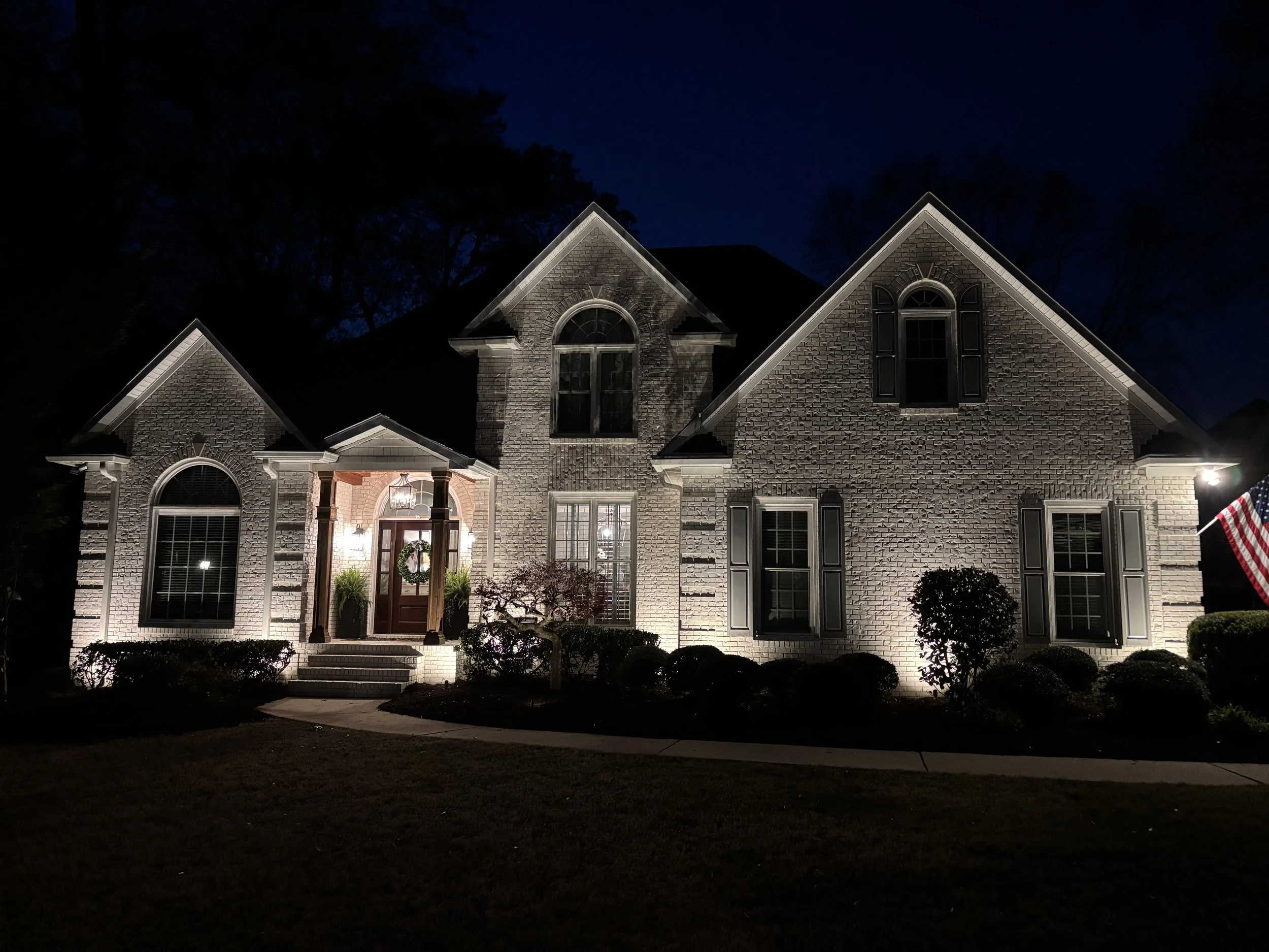 Illuminated brick house at night with American flag, front steps, porch with lanterns, bushes, and trees in background.