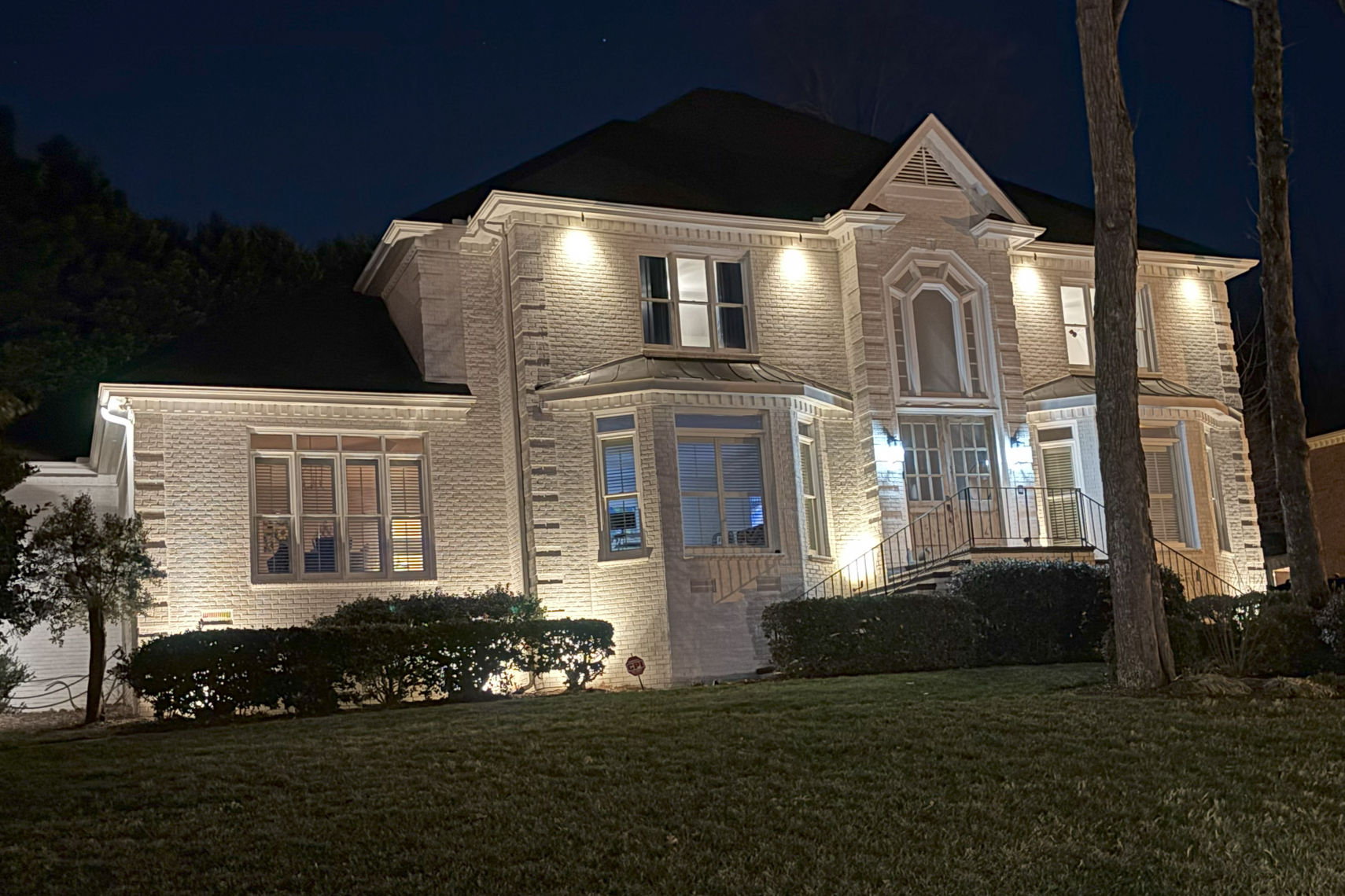 A lit up two-story house at night with exterior lights, a staircase leading up to the front door, brick walls, multiple windows, and a well-maintained lawn with trees and shrubs.