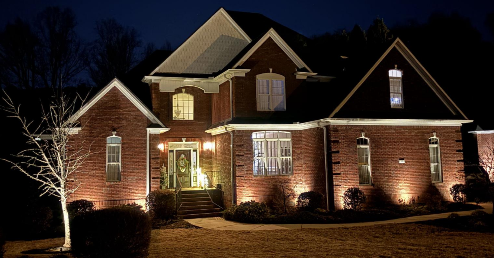 A large brick house illuminated at night, with a front door decorated with a holiday wreath, surrounded by bushes and trees, with a clear pathway leading to stairs.