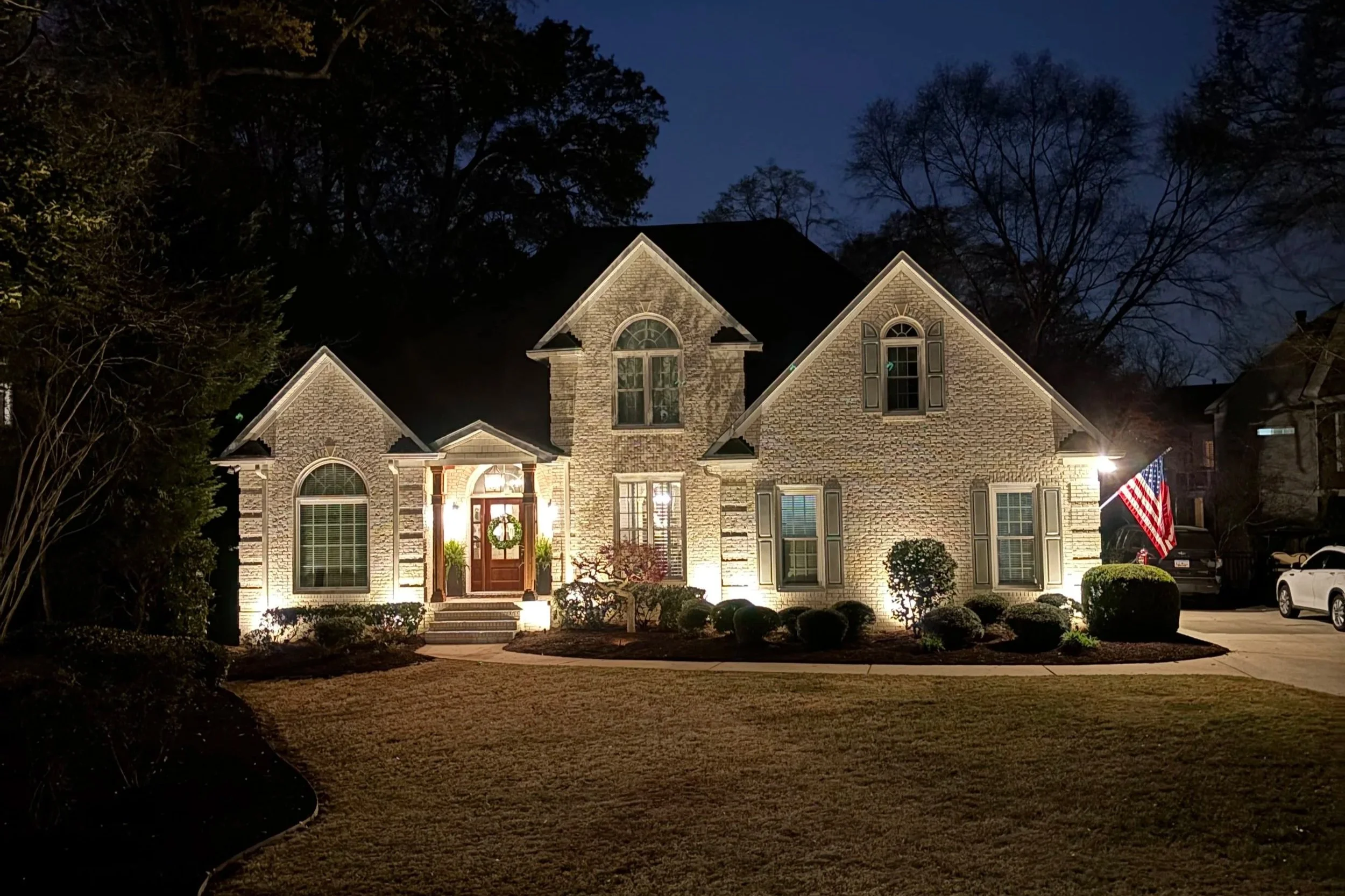 A well-lit brick house at night with a front yard, American flag, and cars parked in the driveway.