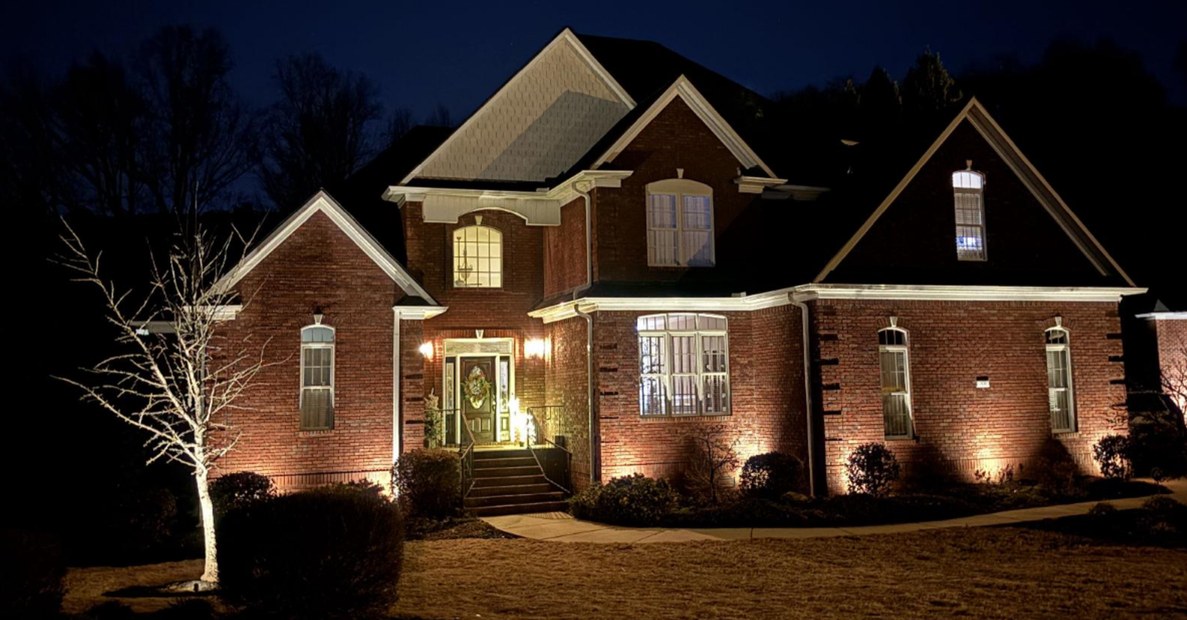 A large brick house illuminated at night, featuring a front porch with steps, multiple windows, and a tree to the left, with a dark sky and trees in the background.